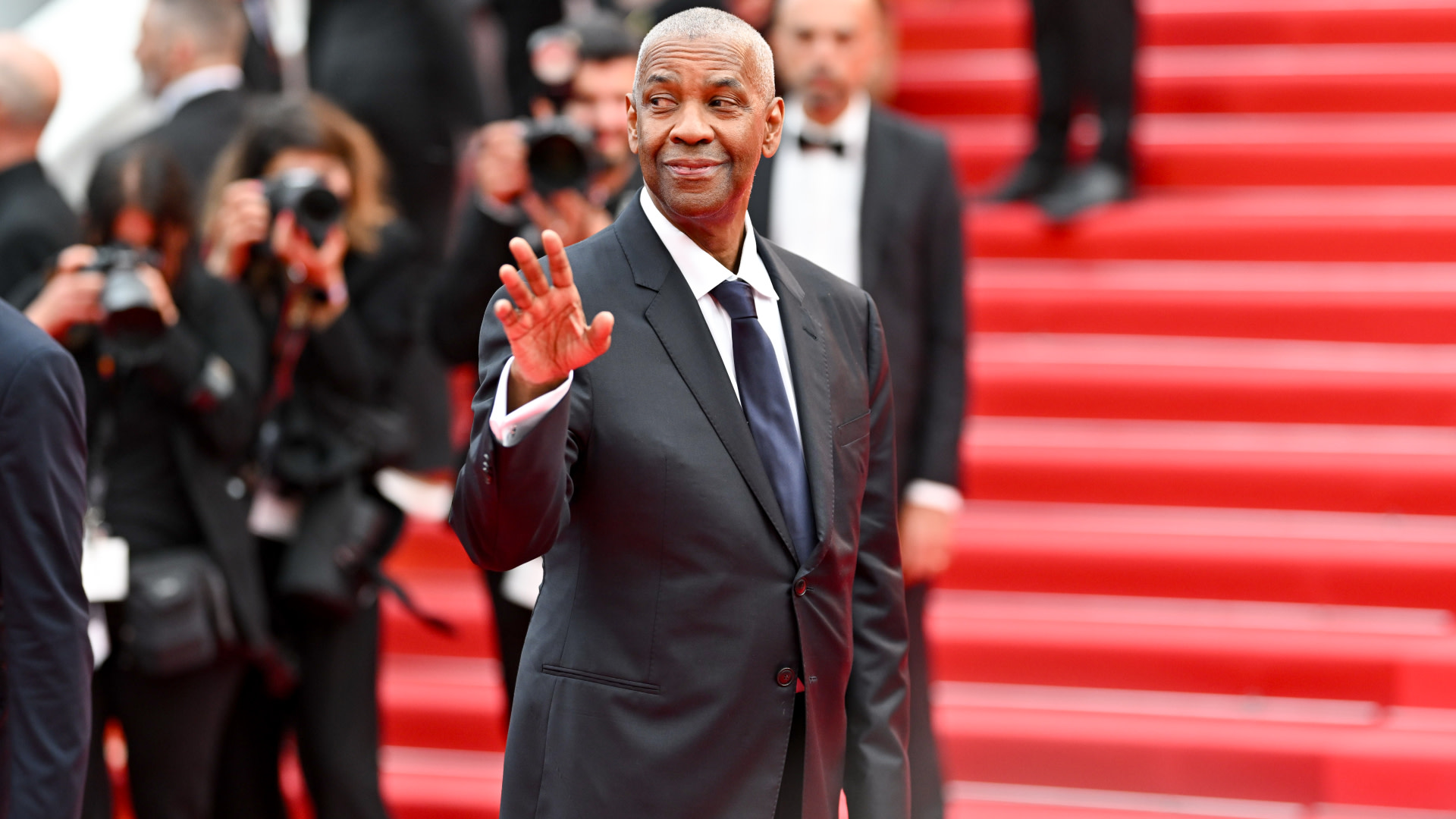 Denzel Washington waves on a red carpet, dressed in a black suit and tie, with photographers in the background.