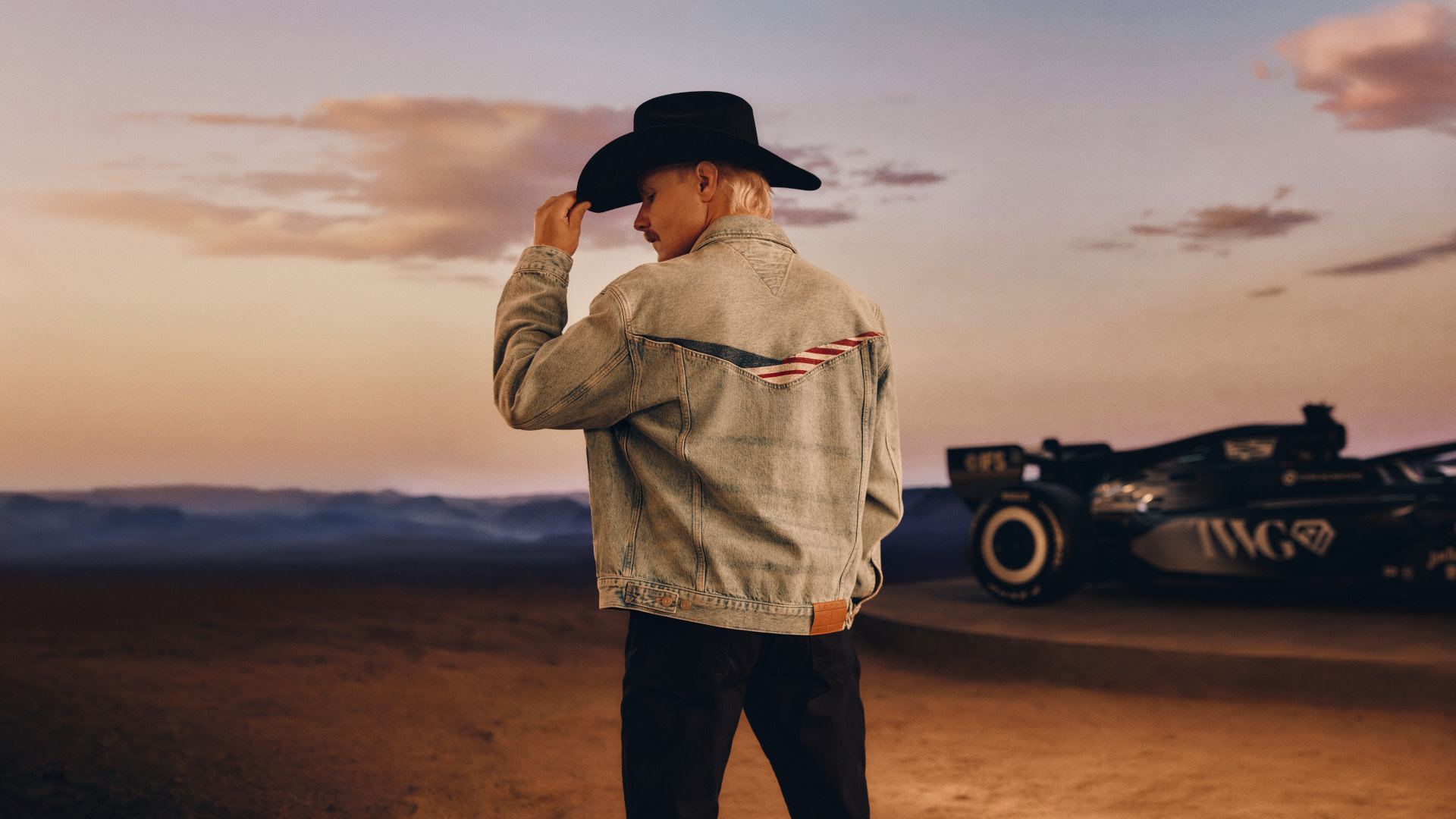 A person in a denim jacket and cowboy hat stands in a desert setting, with a race car in the background at sunset.