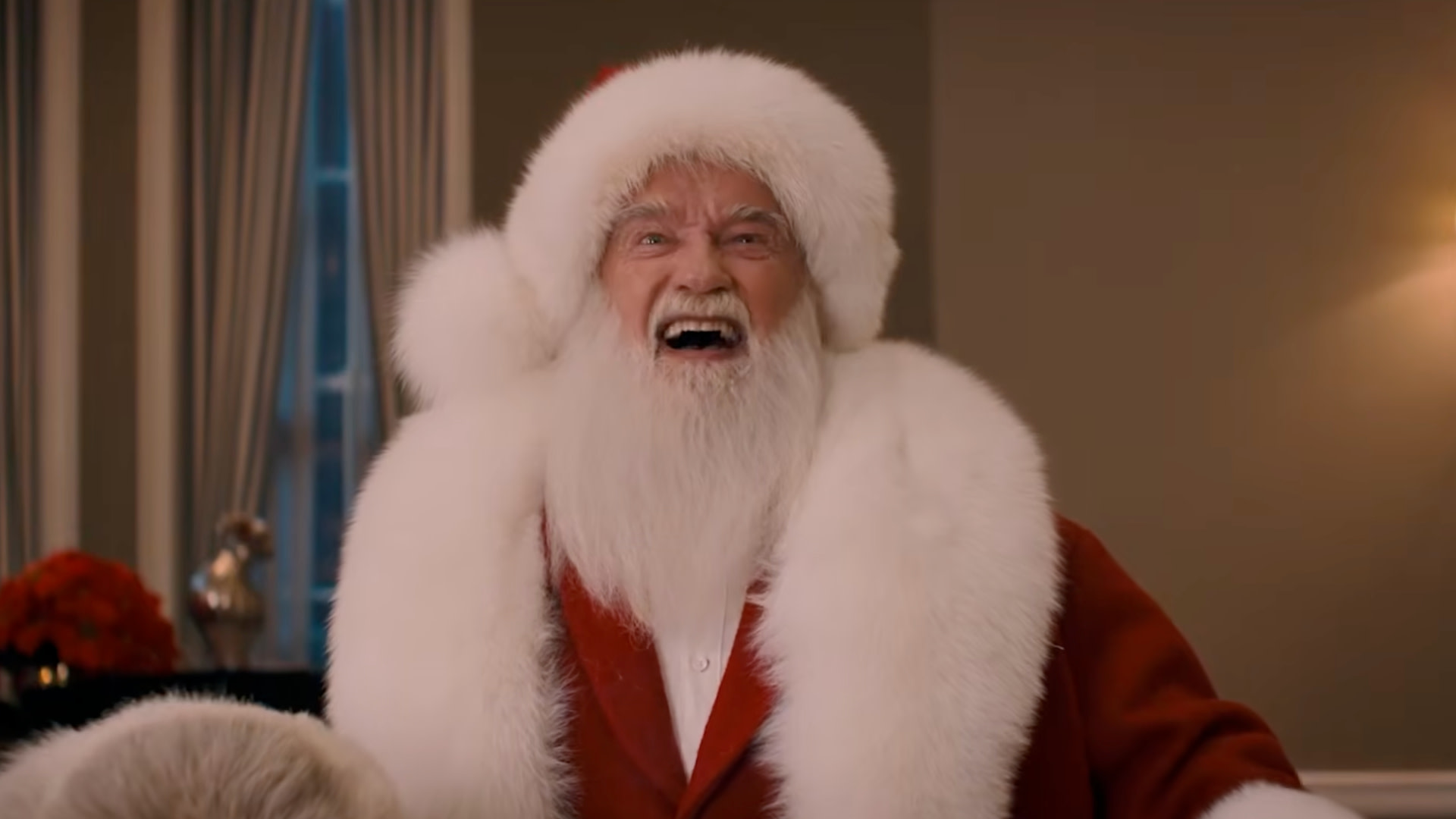 Arnold Schwarzenegger dressed as Santa Claus with a red suit, white beard, and fur trim, smiling broadly in a warmly lit room.