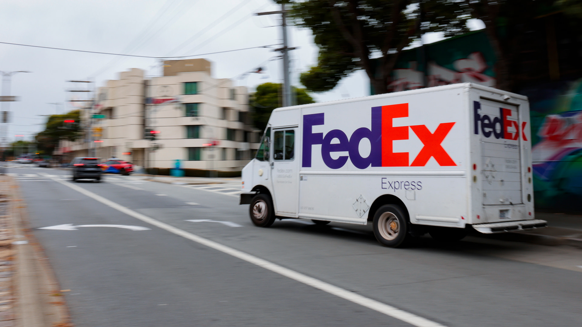 A FedEx delivery truck driving down a city street, with buildings and trees in the background.