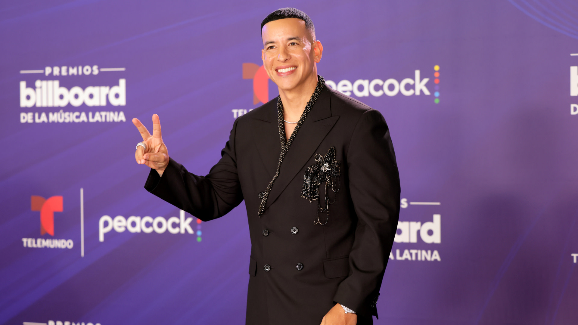 Daddy Yankee at the Billboard Latin Music Awards, wearing a black suit with a beaded design, smiling and making a peace sign.