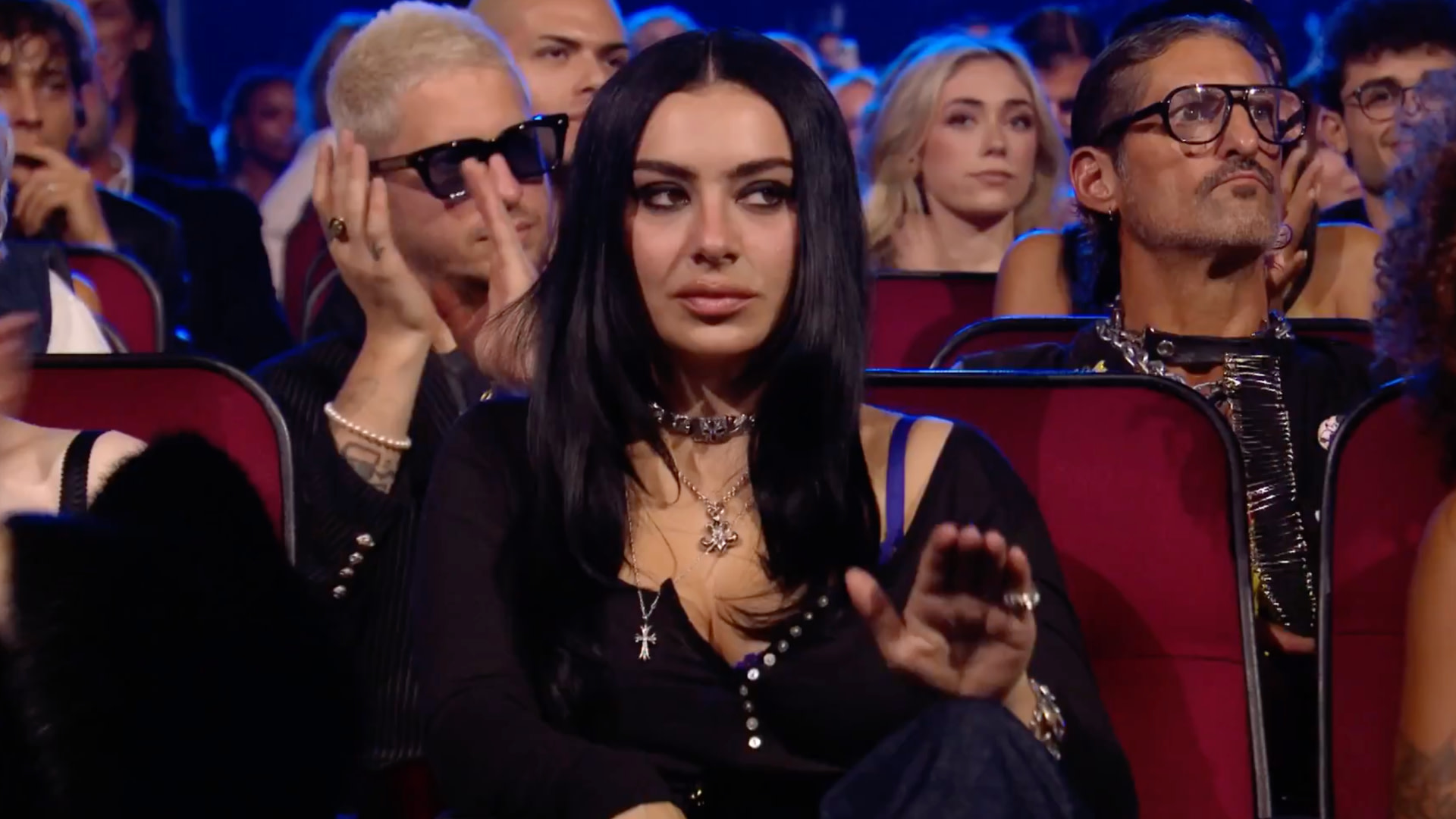 People sitting in an audience, with Charli xcx in the center wearing black, surrounded by others in stylish attire, clapping.
