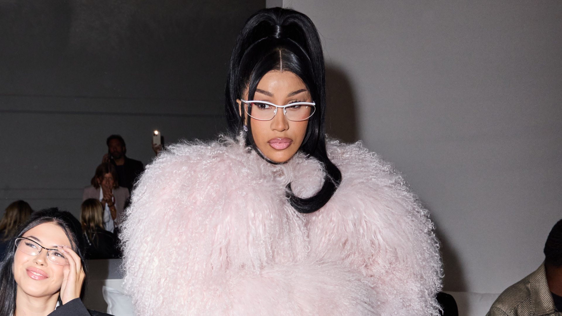 Cardi B in a fluffy pink outfit and glasses, with a high ponytail, at a fashion event.