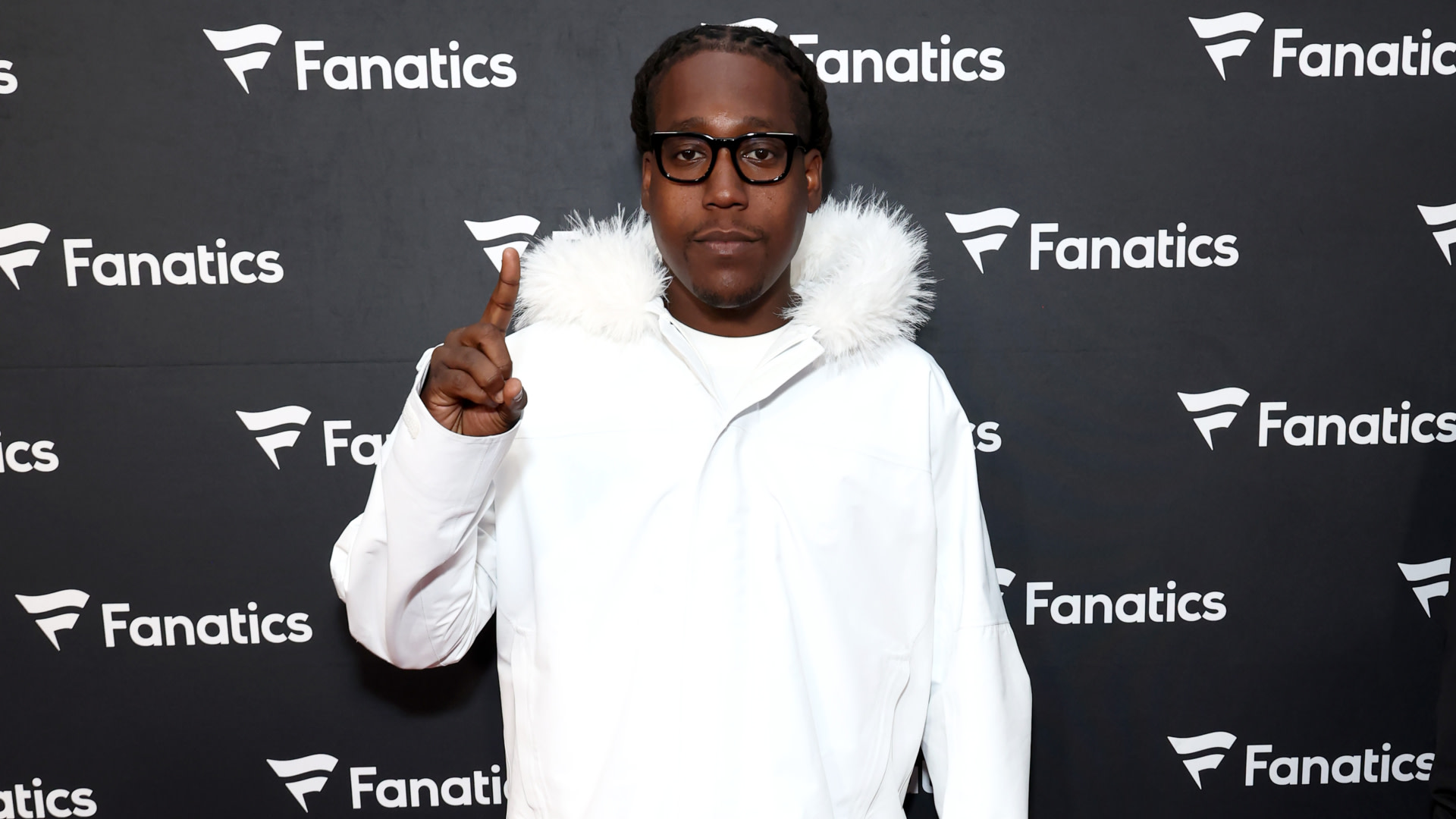 Don Toliver in a white jacket and glasses poses on the red carpet at a Fanatics event, making a "number one" gesture.