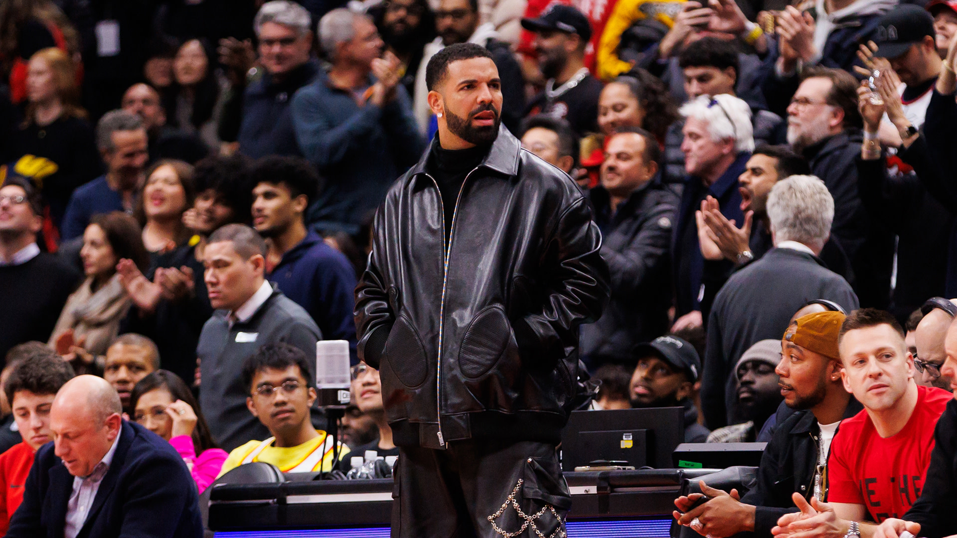 Drake at a basketball game, wearing a black leather jacket, surrounded by a crowd.