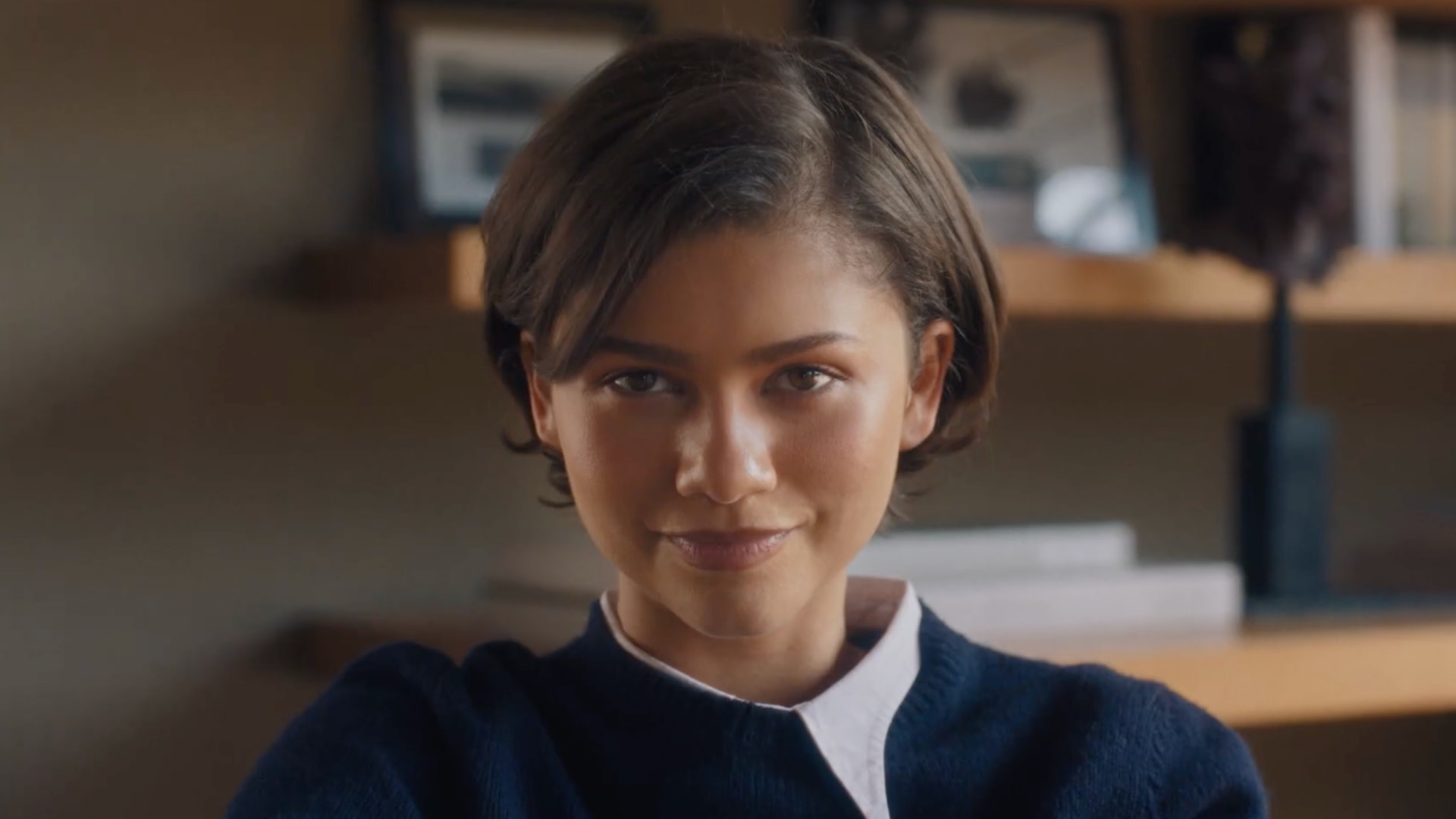 Zendaya with short hair, wearing a navy blue sweater over a white shirt, smiling slightly, with shelves and framed photos in the background.