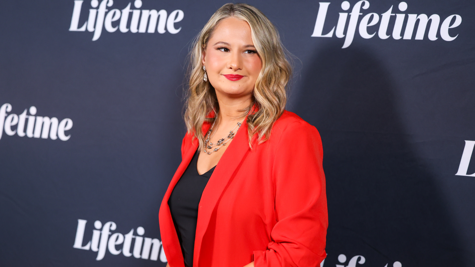 Gypsy Rose Blanchard in a red blazer poses on the red carpet in front of a "Lifetime" backdrop.