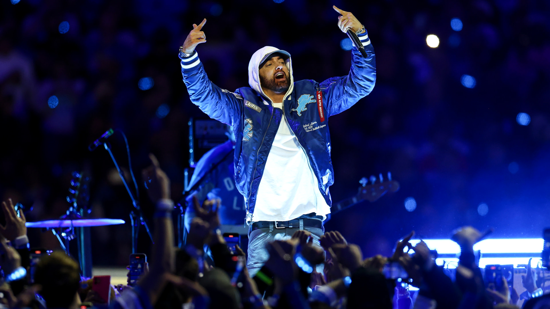 Eminem in a blue jacket and white hoodie energetically gestures on stage, surrounded by a cheering crowd in a brightly lit arena.