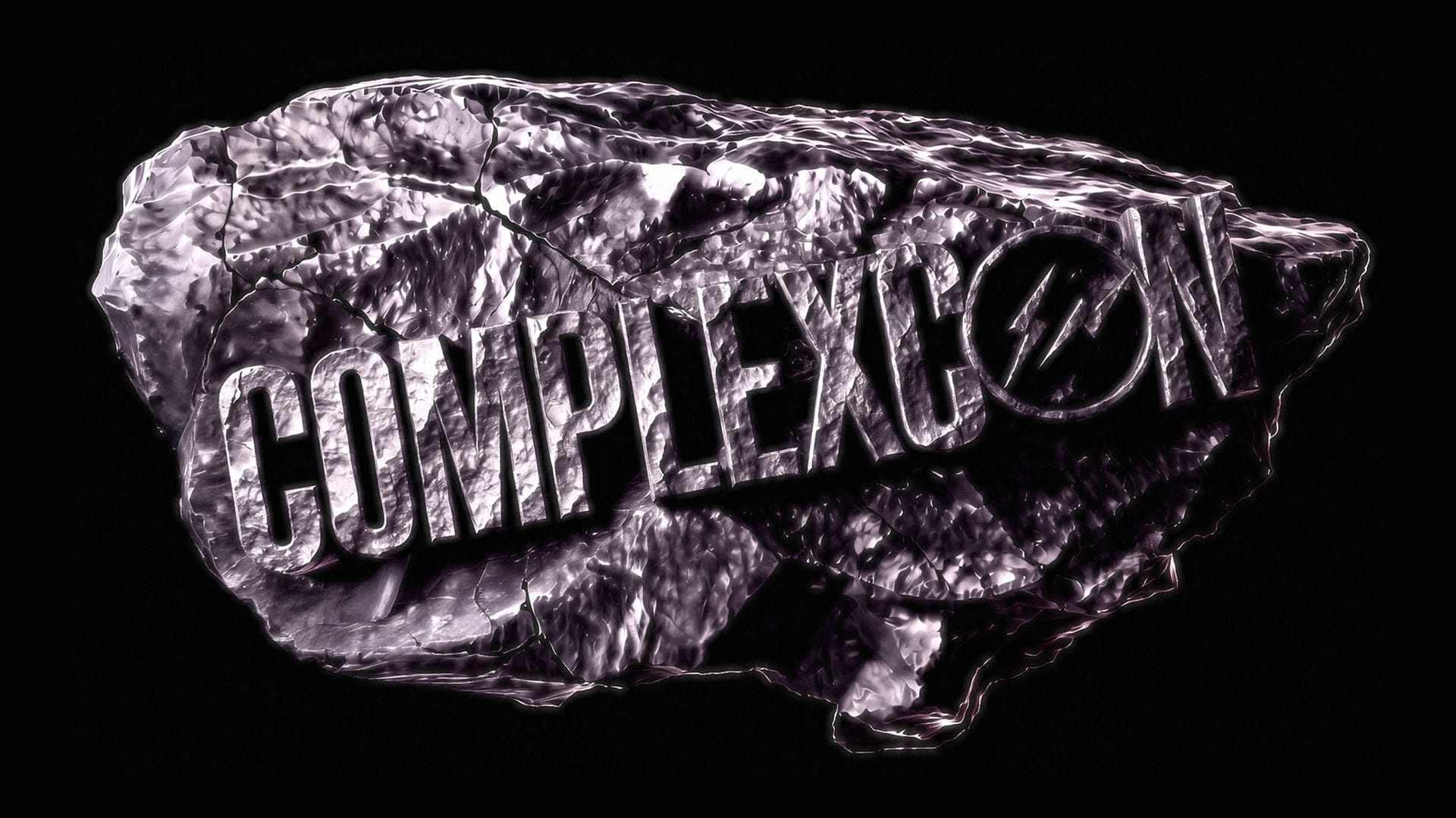 Text "COMPLEXCON" on a textured, metallic rock surface with a lightning bolt in the letter "O," set against a black background.
