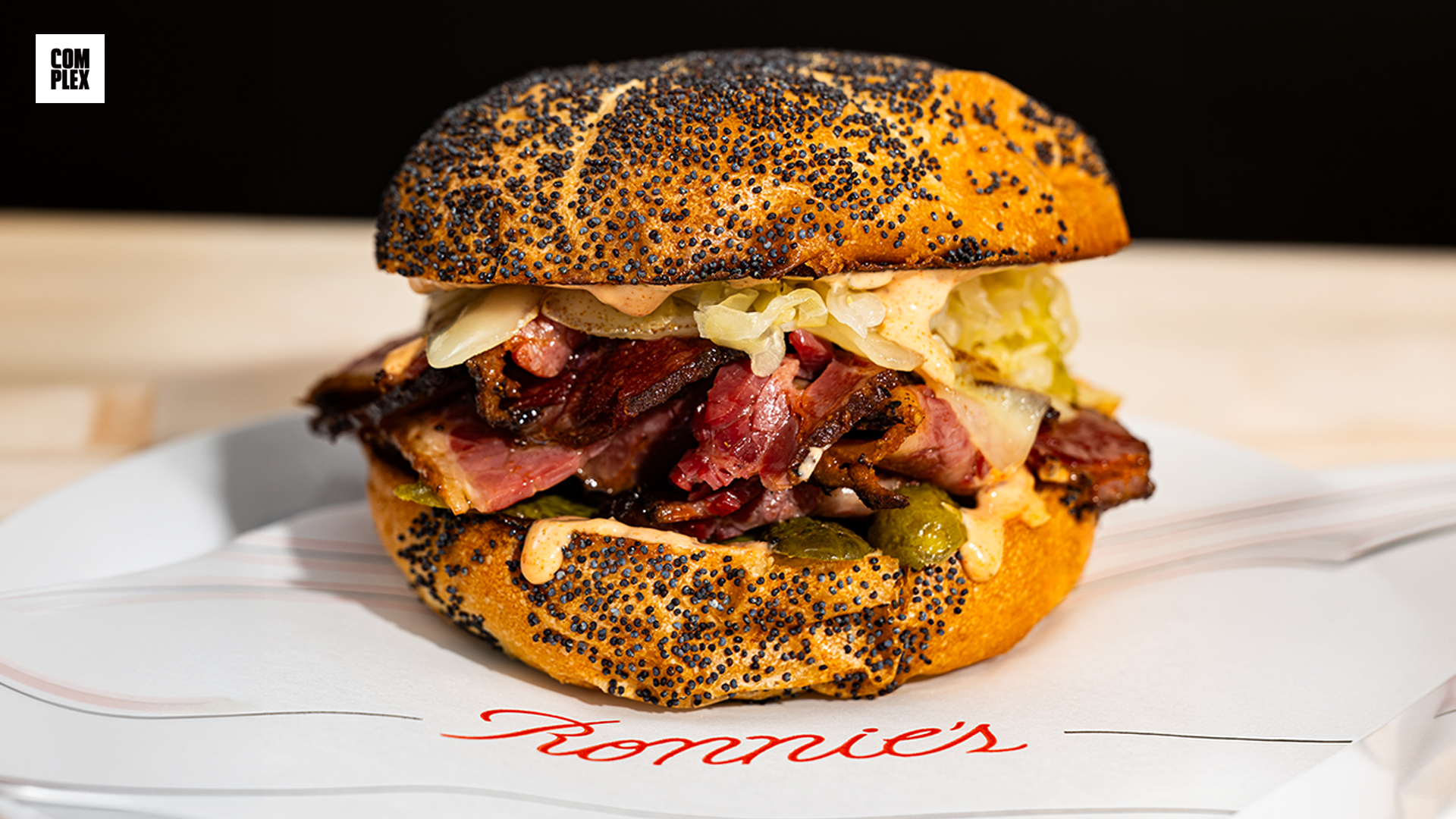 A poppy seed bun sandwich filled with pastrami, sauerkraut, and pickles, placed on a napkin with "Ronnie's" written on it.