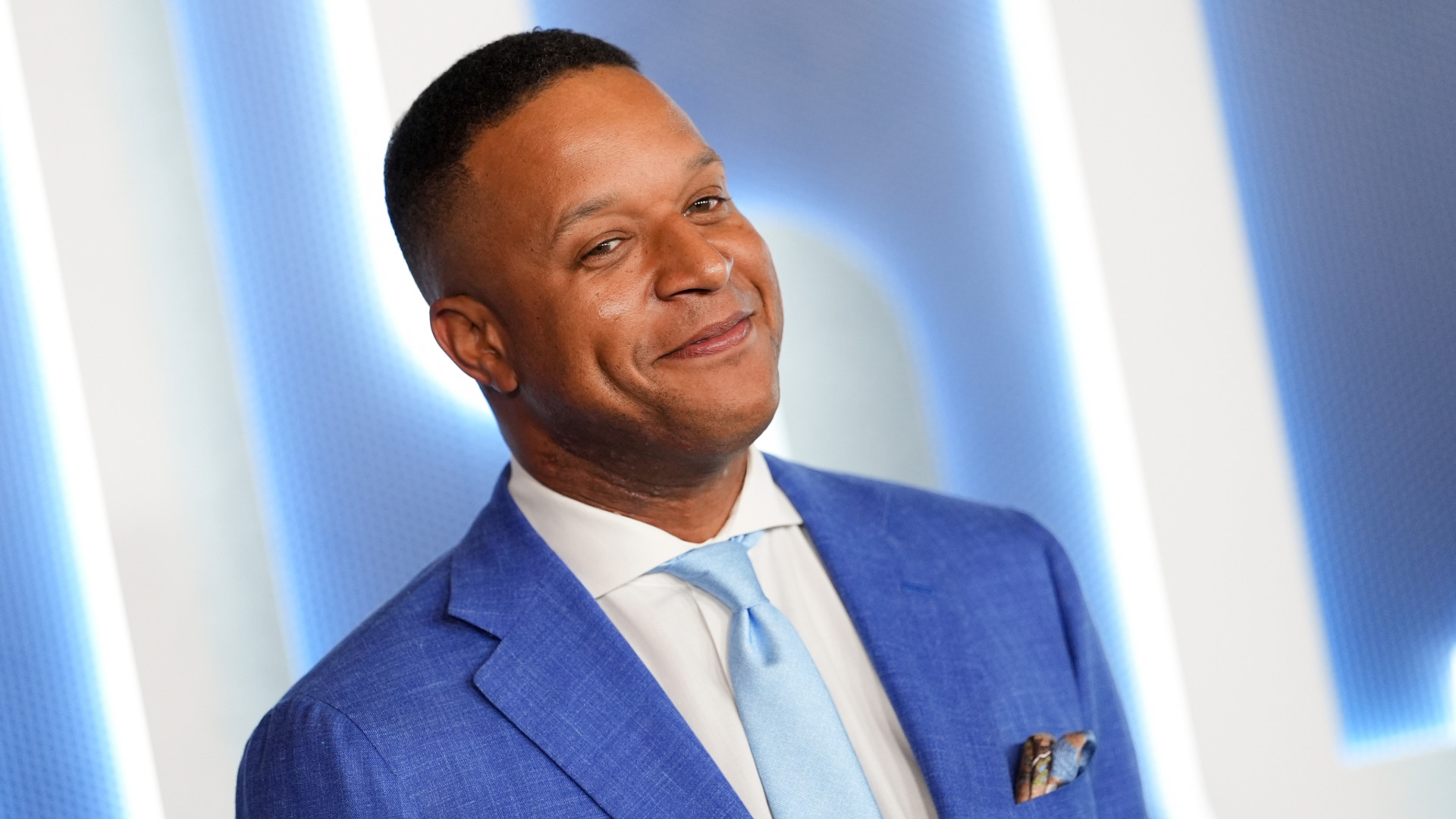 Man in a blue suit and light blue tie smiling against a bright background.