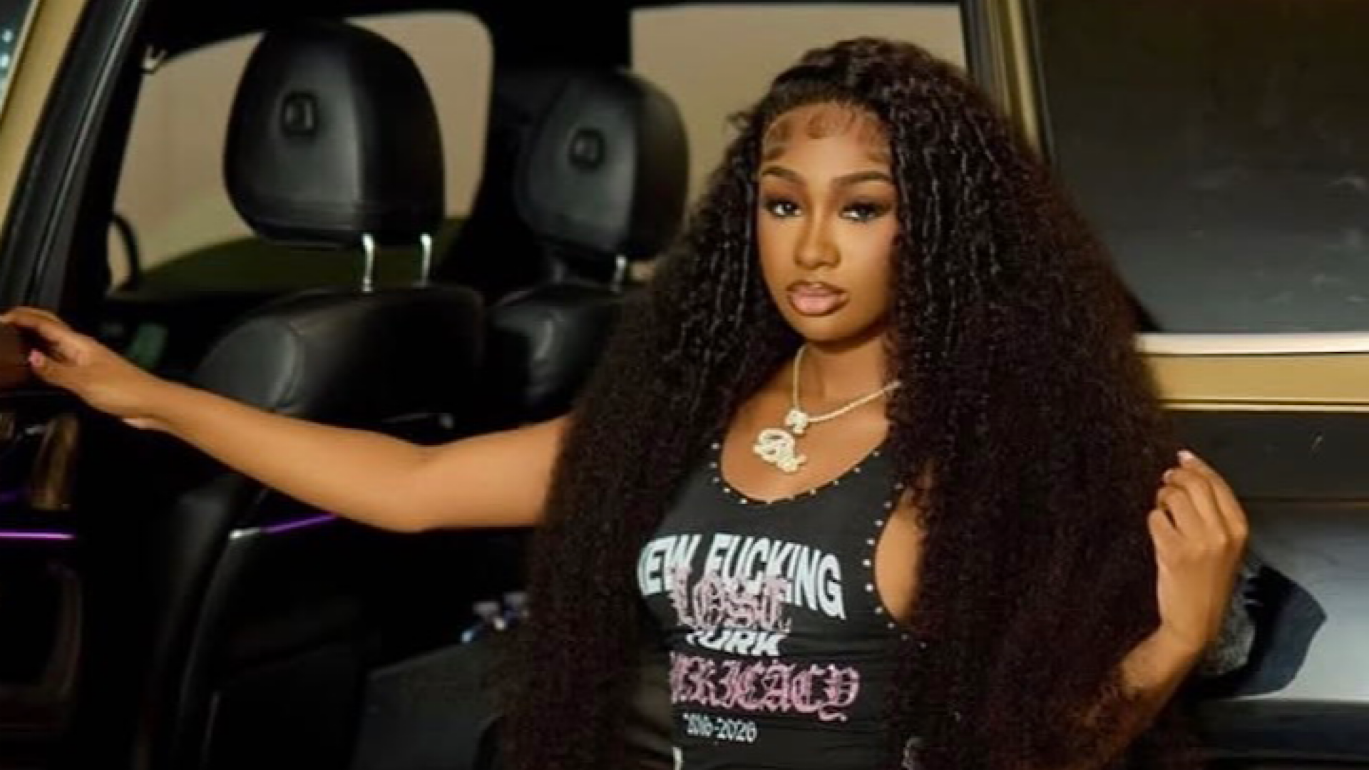 A woman with long curly hair poses confidently next to a car, wearing a black top with text and a necklace.
