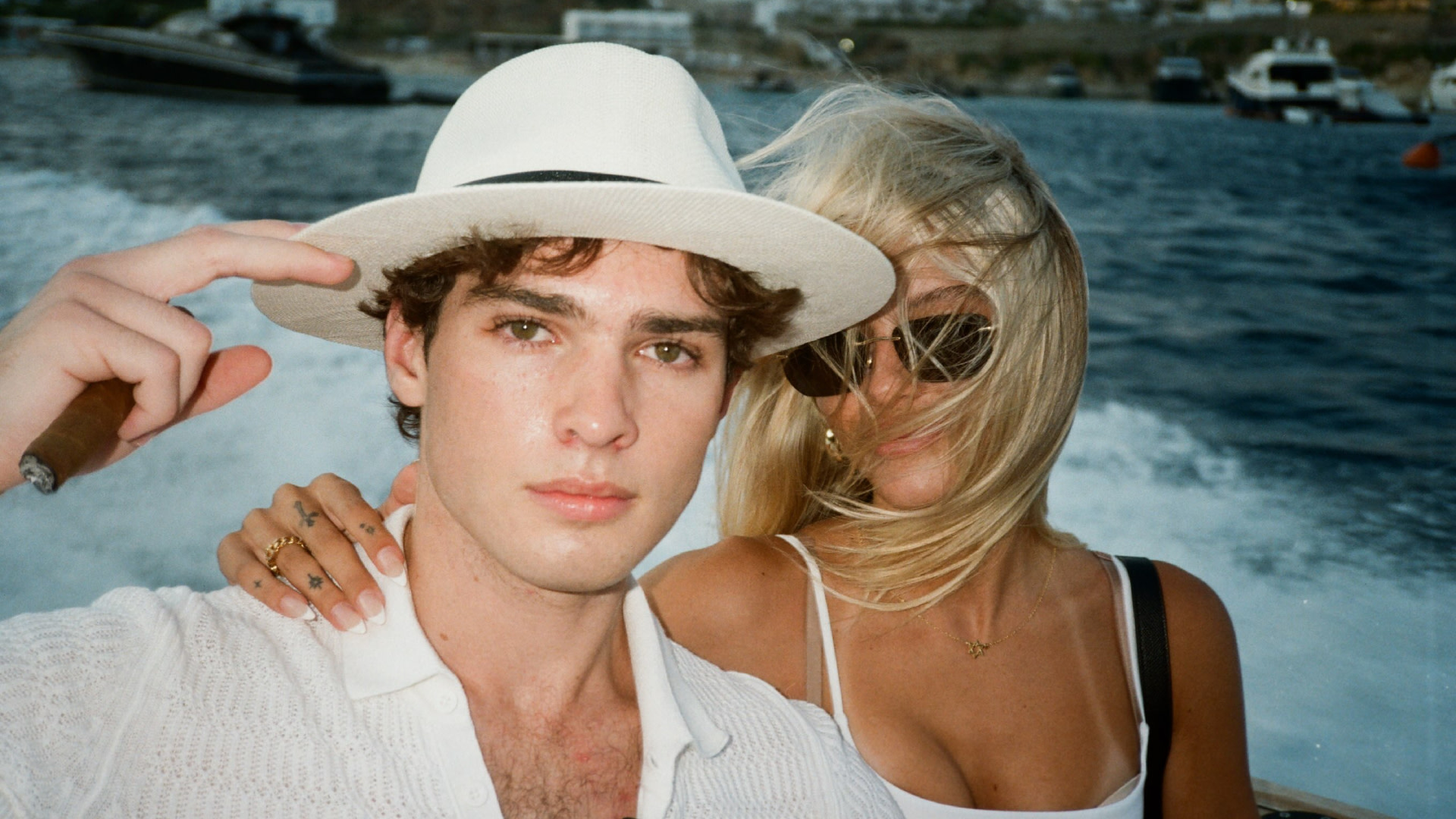 Clavicular in a white hat and shirt poses with girlfriend Lily Snyder in sunglasses on a boat, with the ocean and yachts in the background.