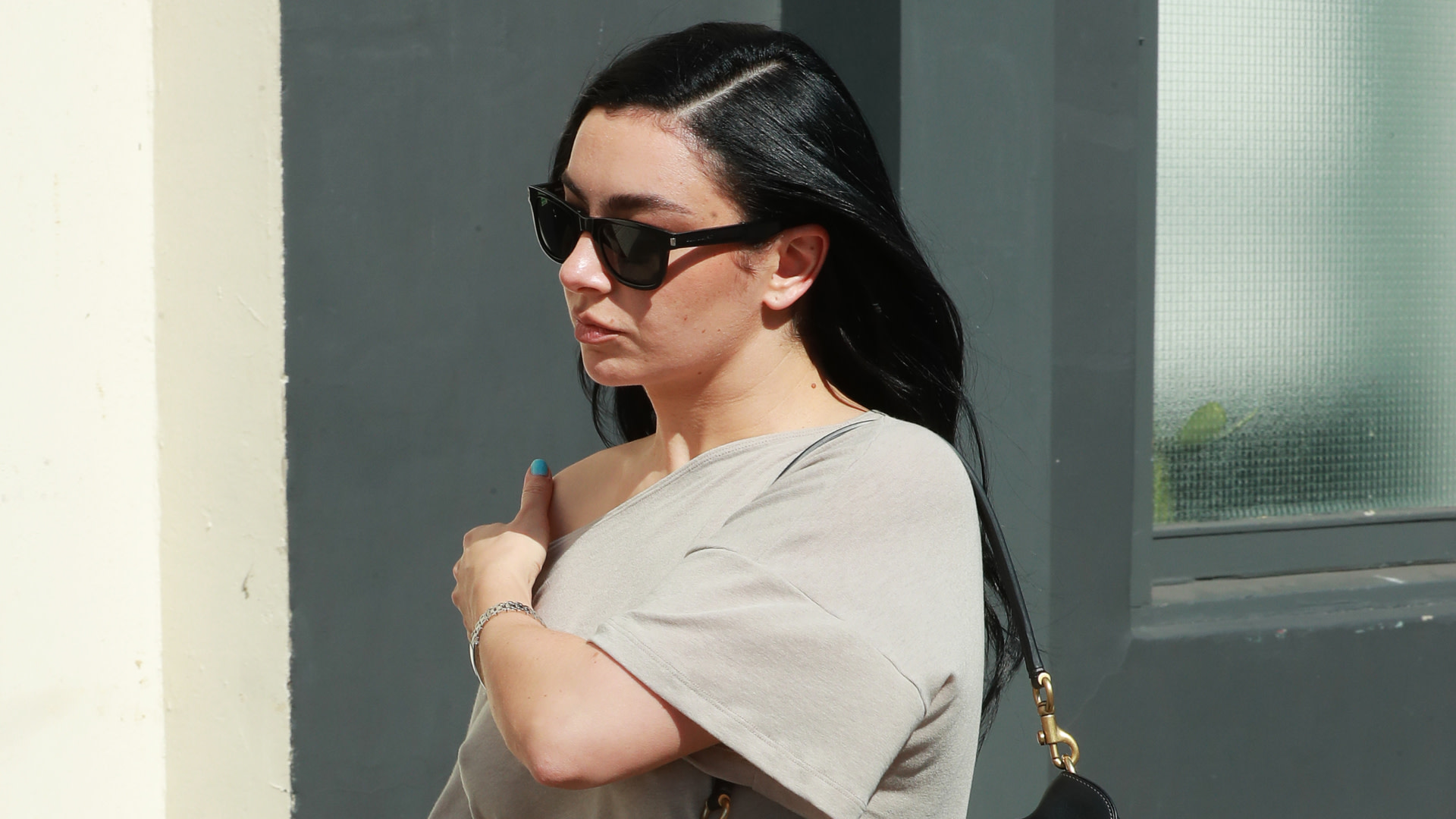 Charli xcx with long black hair wearing sunglasses and a gray top stands outside near a building.