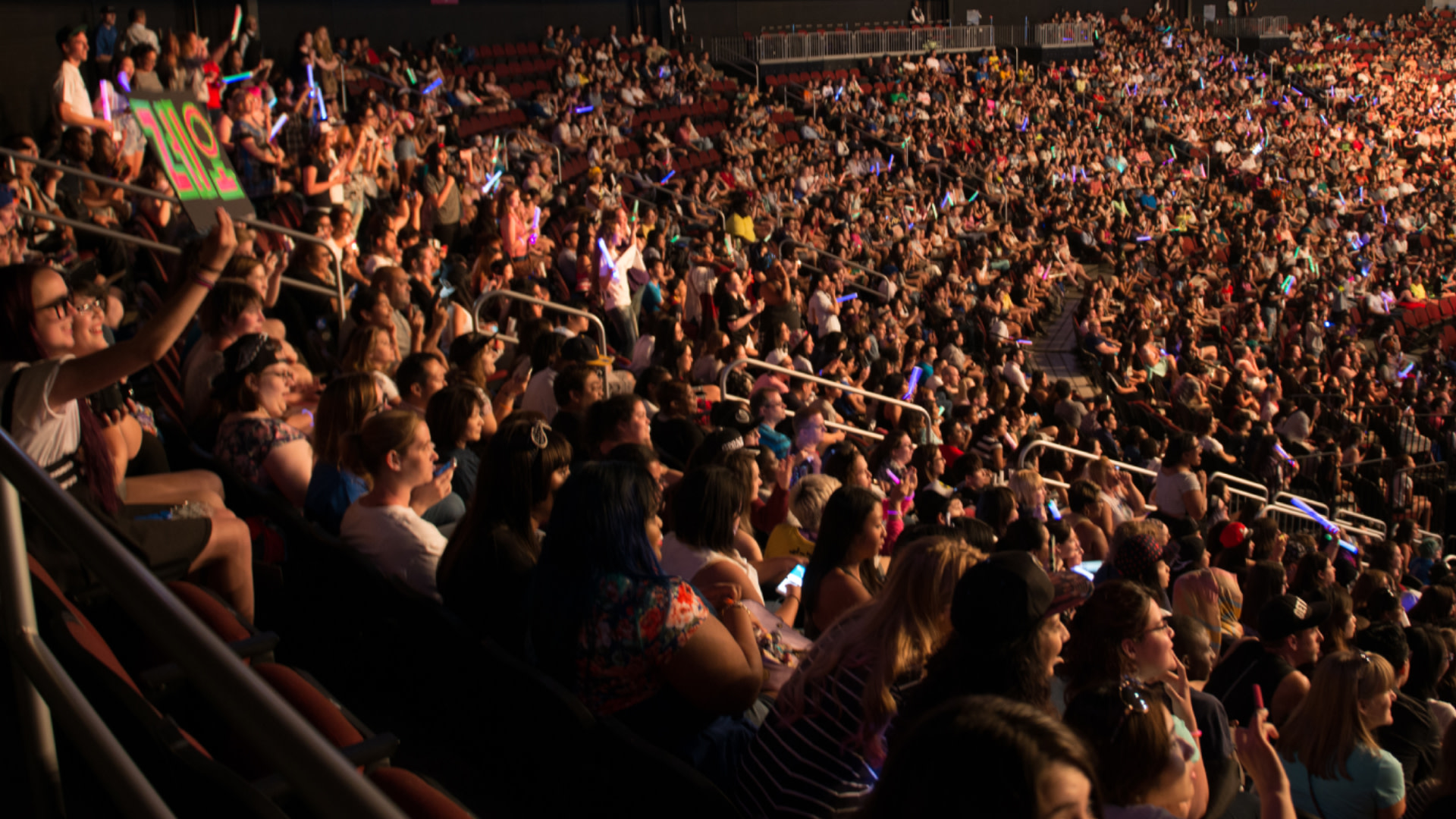 A large crowd in an arena, cheering and holding signs, with colorful lights visible.