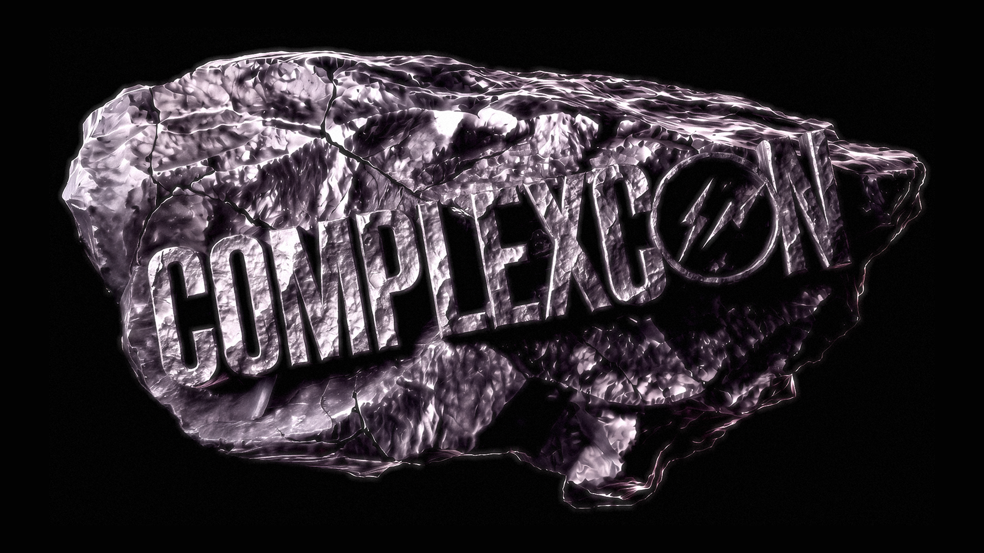 Text "COMPLEXCON" on a textured, metallic rock surface with a lightning bolt in the letter "O," set against a black background.