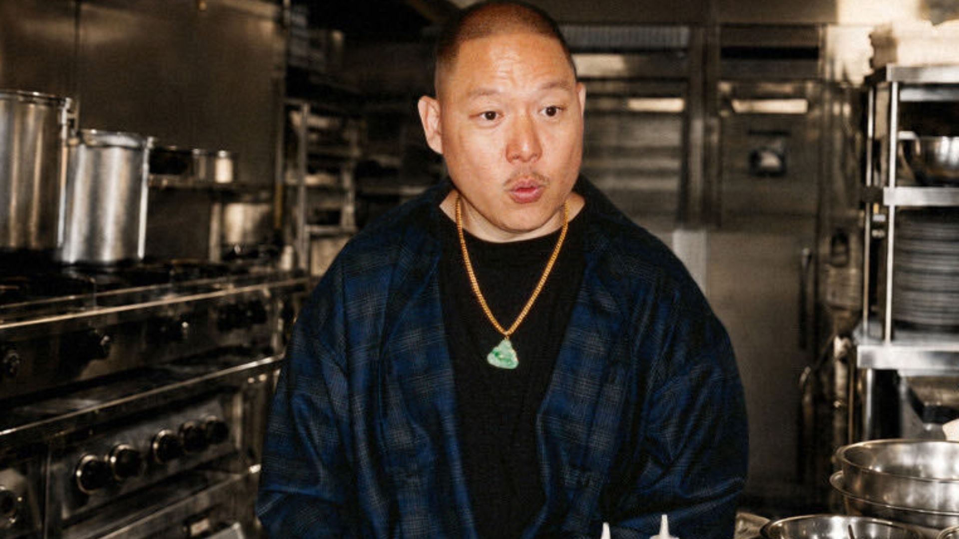Eddie Huang stands in a kitchen, wearing a dark plaid jacket and a jade necklace. Stainless steel kitchen equipment surrounds him.
