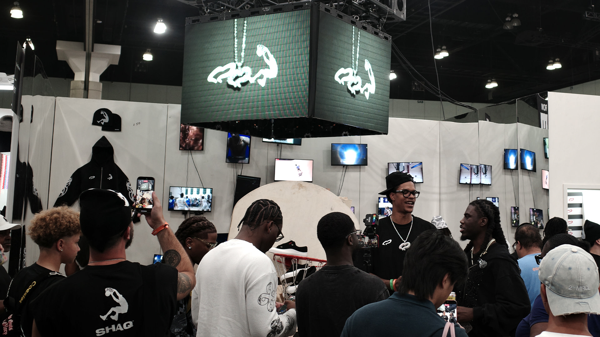 A crowded booth at a convention with people gathered around, some wearing branded clothing. Monitors and a logo are displayed above.