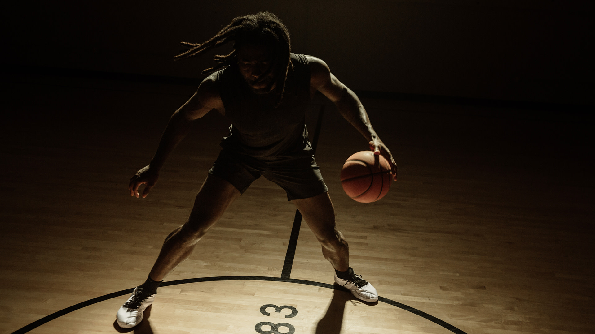 A basketball player dribbling on a dimly lit court. The player wears dark athletic clothing and has dreadlocks.