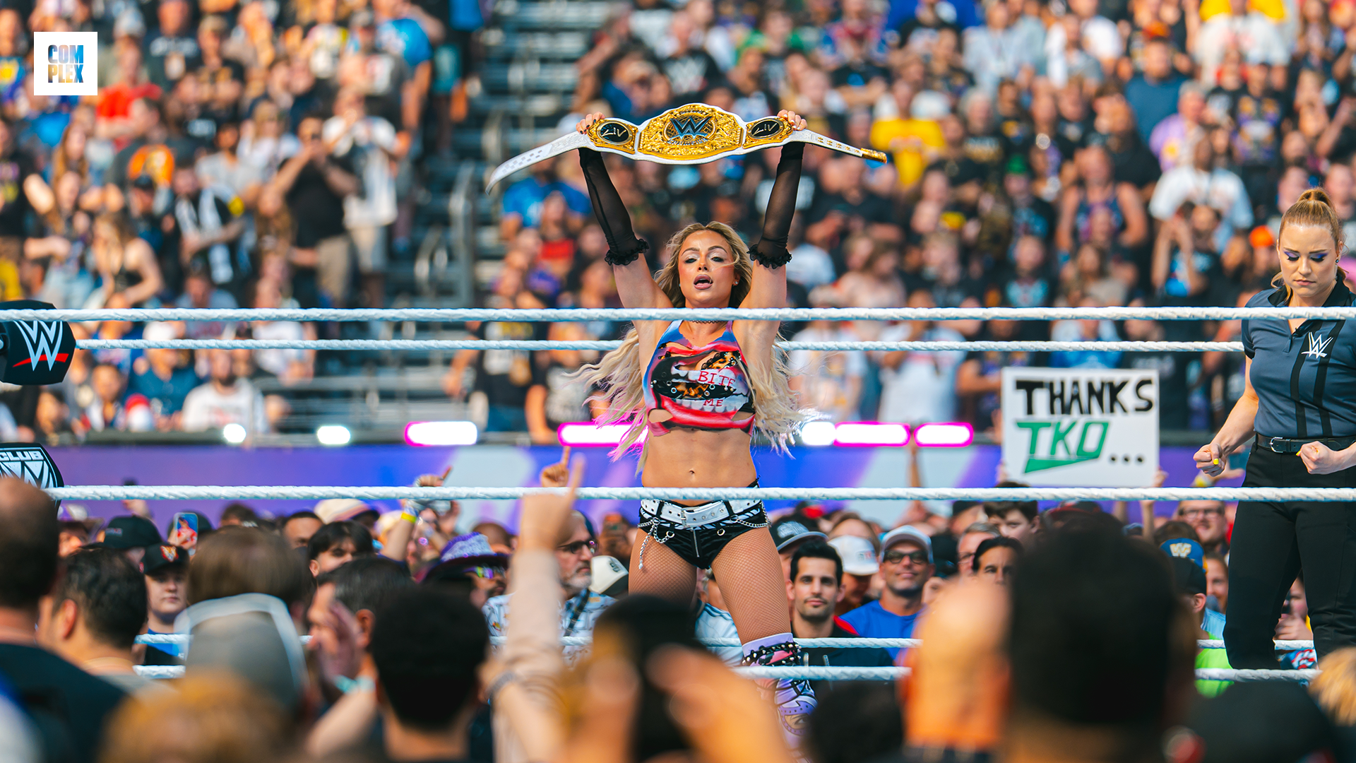 A female wrestler holds a championship belt in a crowded wrestling arena, celebrating her victory.