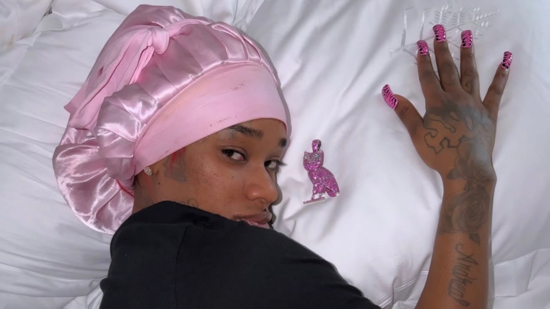 Sexyy Red with tattoos and pink bonnet lying on a white pillow, wearing pink patterned nails and a small pink OVO figurine nearby.
