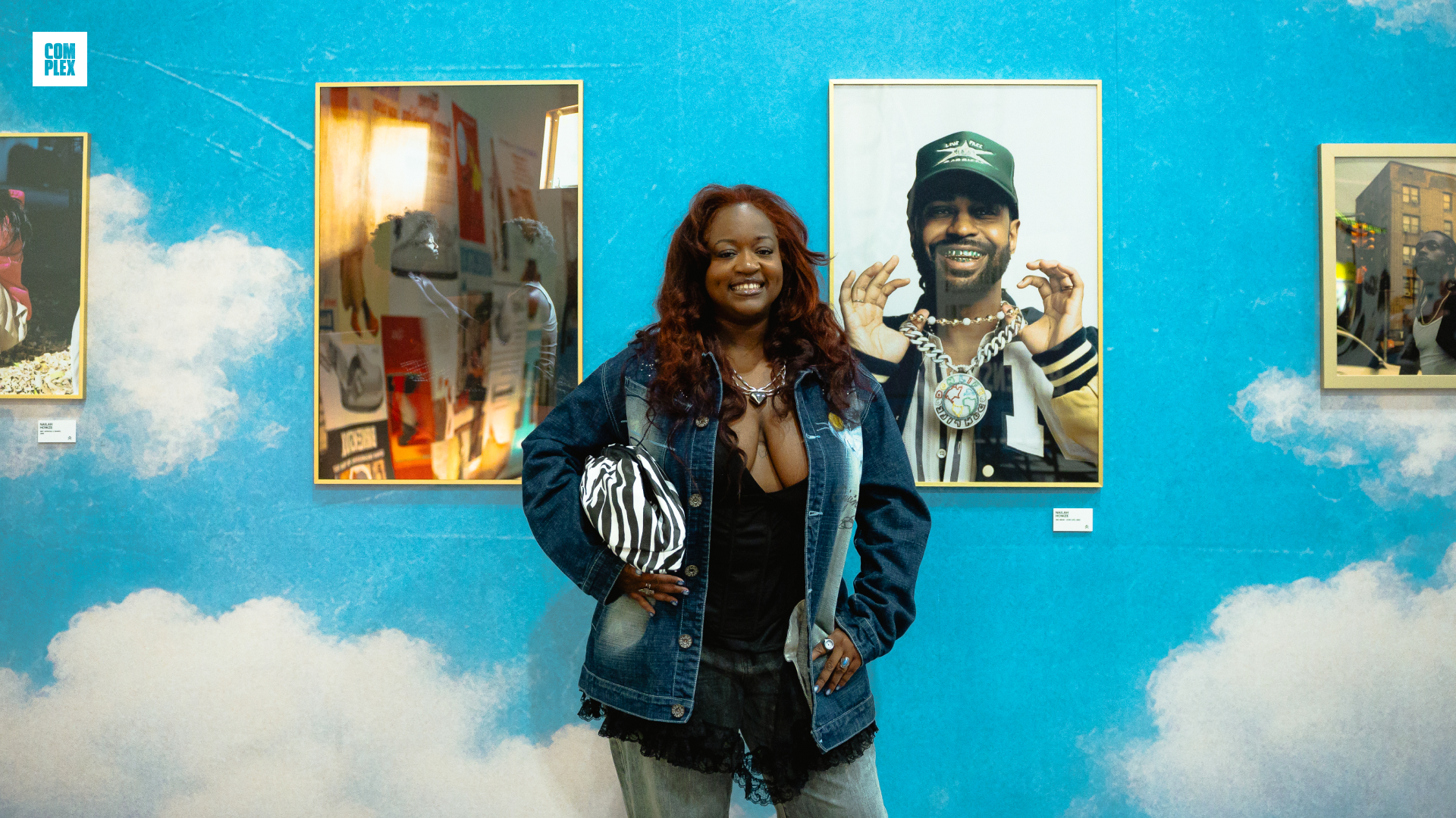 A woman smiles in front of a blue wall with cloud patterns, featuring framed photographs. She wears a denim jacket and holds a striped bag.