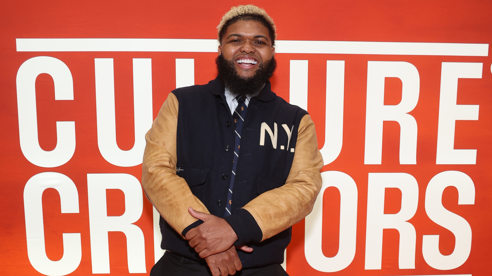 Druski with a beard and blonde-tipped hair, wearing a jacket with "N.Y." on it, stands smiling in front of a "Culture Creators" sign.
