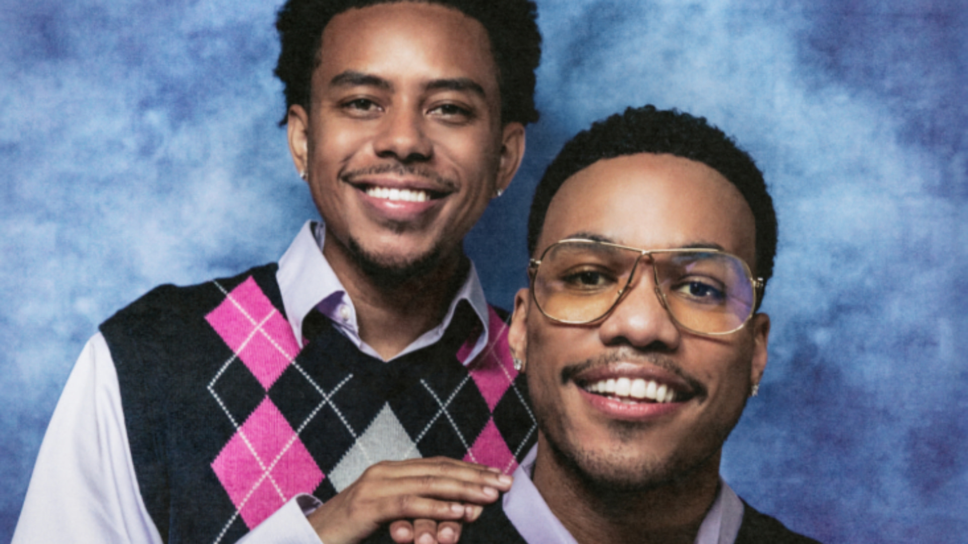 Cordae and Anderson .Paak smiling men in argyle vests pose against a blue backdrop; one wears glasses.