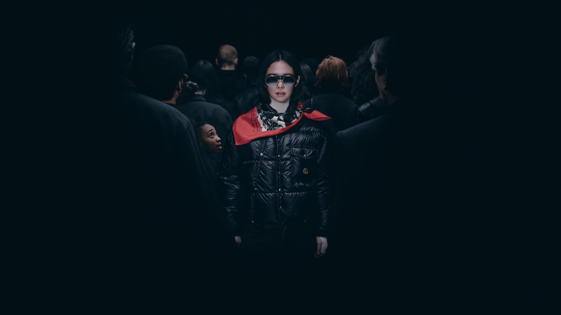 Jennie in a stylish black and red coat and sunglasses stands out in a crowd, illuminated against a dark background.