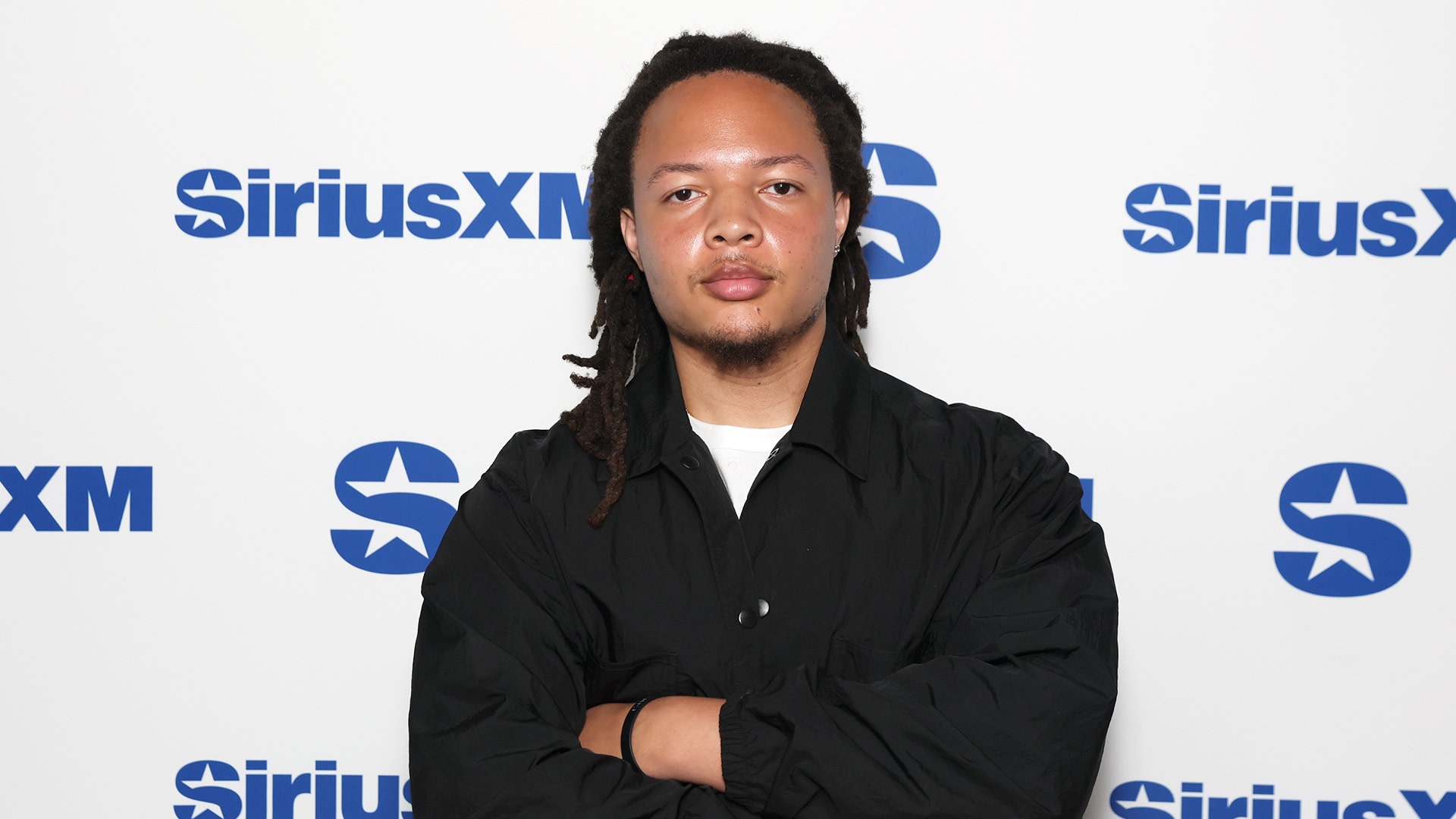 A person with dreadlocks in a black jacket poses with crossed arms in front of a SiriusXM backdrop.