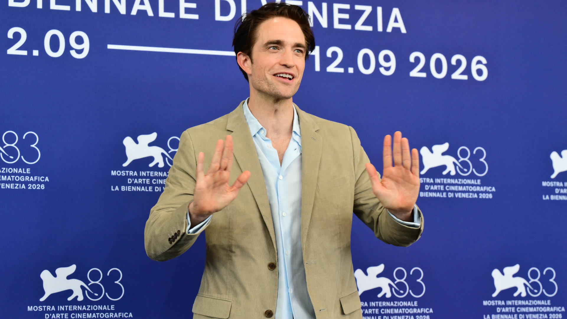Robert Pattinson at the Venice Film Festival, smiling in a beige blazer and light blue shirt, gesturing with hands raised.