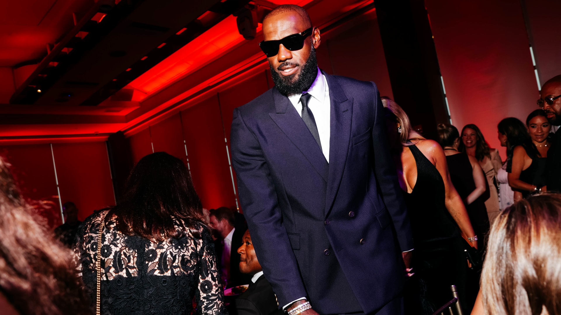 LeBron James in a dark suit and sunglasses at a formal event with red lighting, surrounded by attendees.