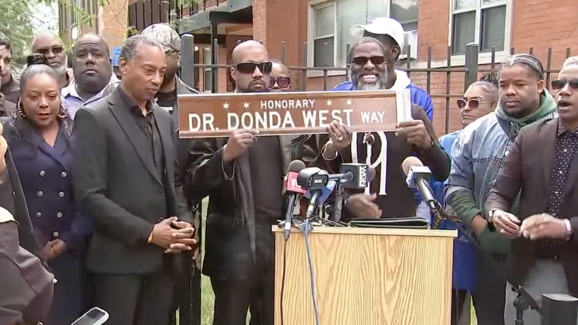 A group of people, including Ye holding a "Dr. Donda West Way" street sign, stand gathered around a podium with microphones.