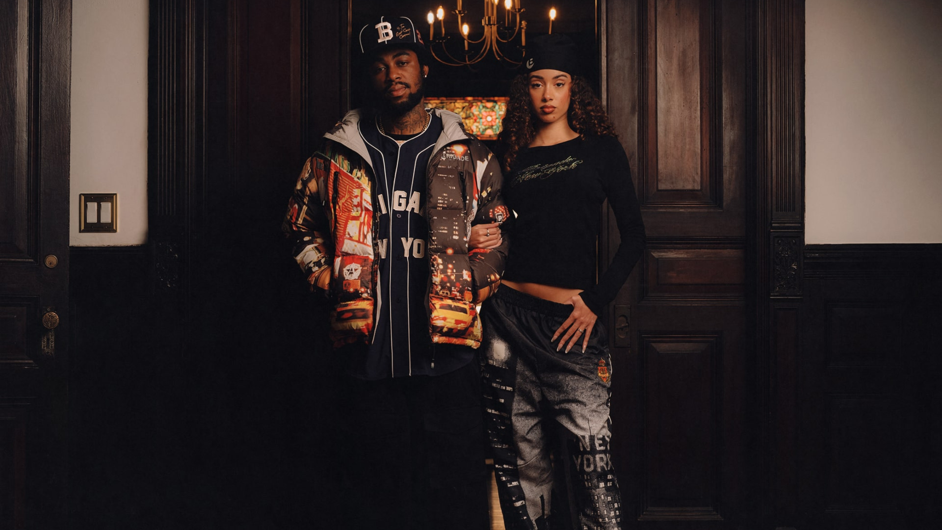 A man and woman in stylish streetwear stand in a dimly lit room with wooden paneling and a chandelier above.