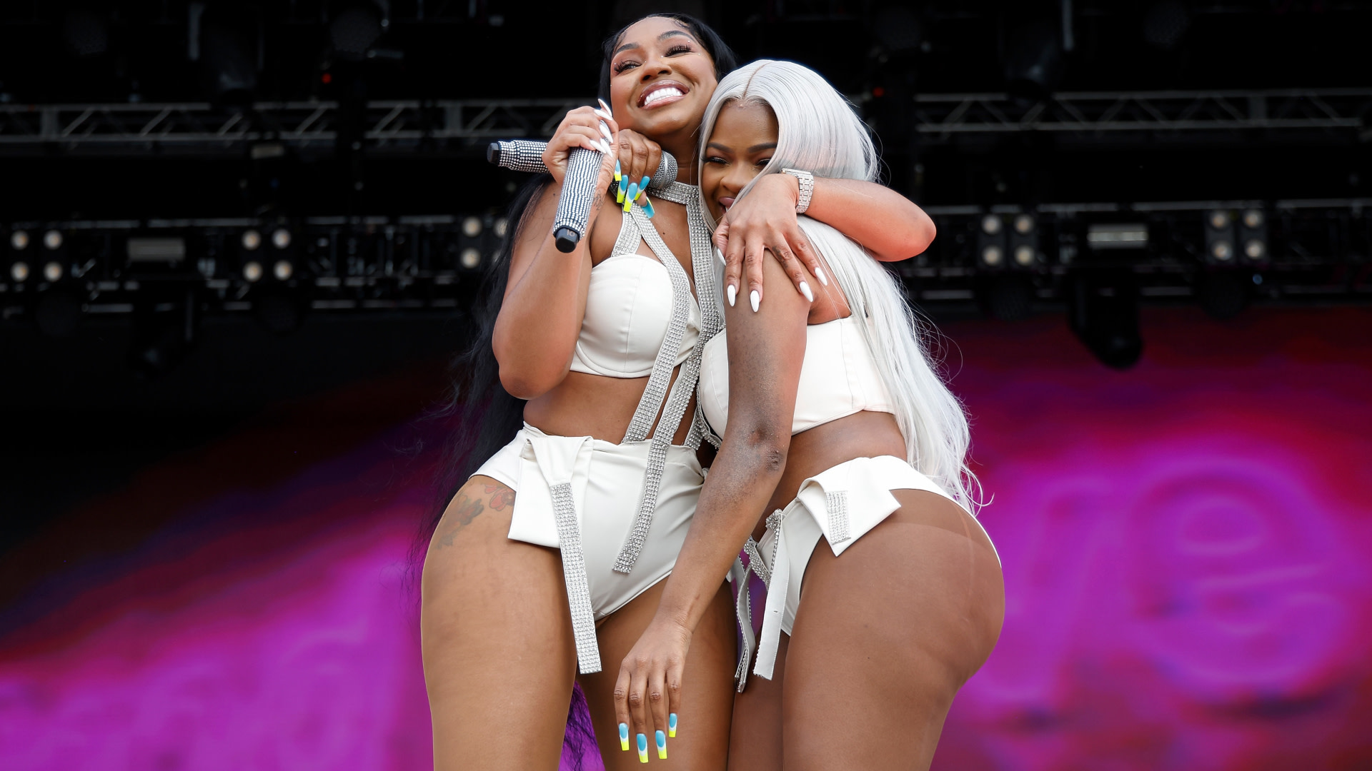 City Girls on stage in matching white outfits, smiling and hugging. One holds a microphone. The background is a vibrant pink.
