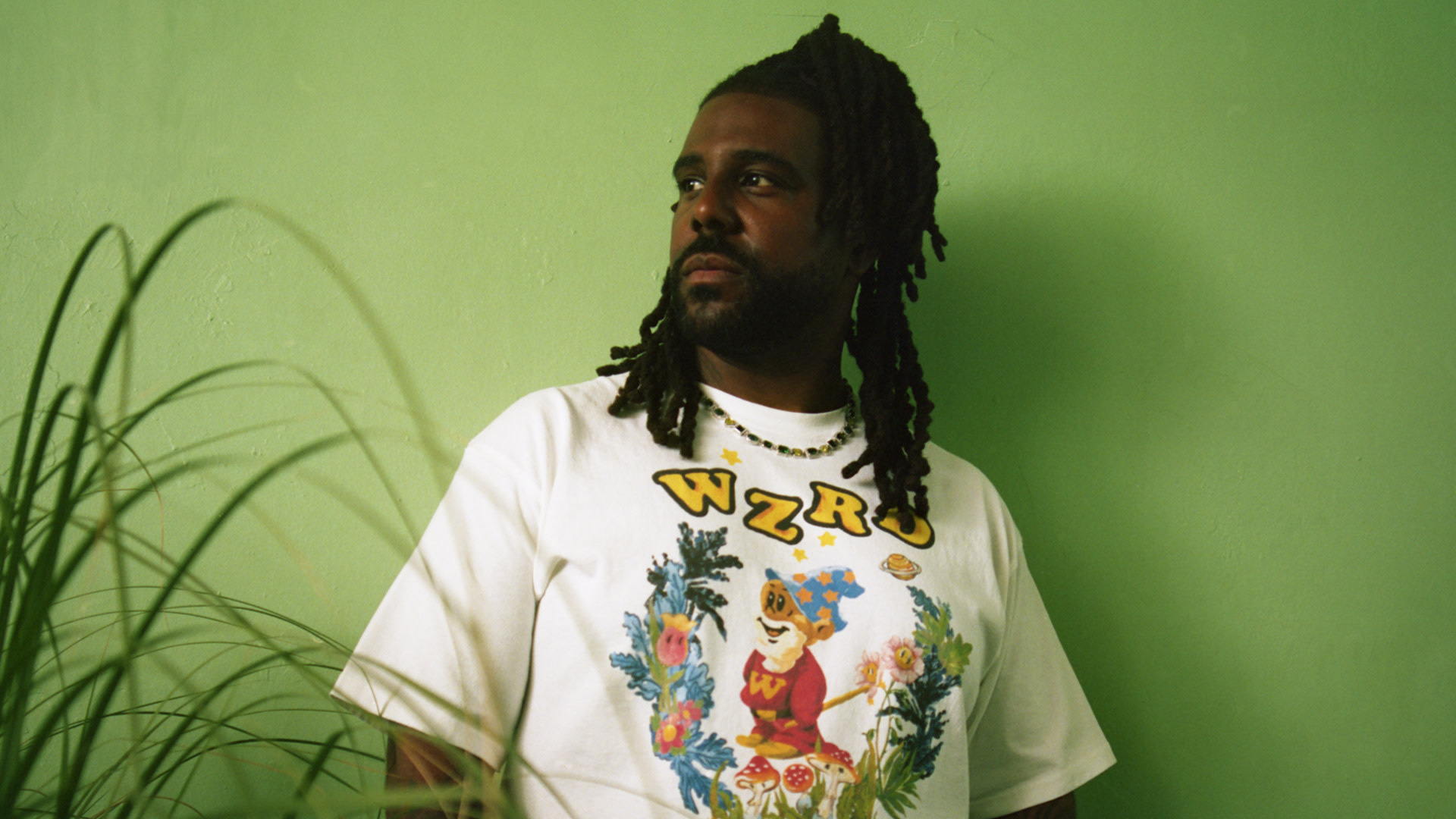 Man with long hair in a white graphic t-shirt, standing against a green wall with plants in the foreground.