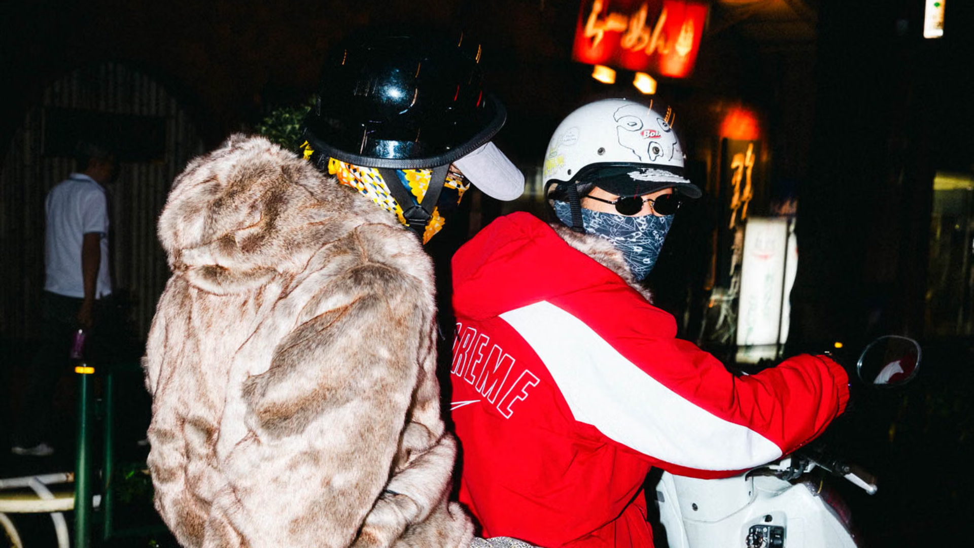 Two people on a scooter at night, wearing helmets and masks. One in a fur coat, the other in a red "Supreme" jacket.
