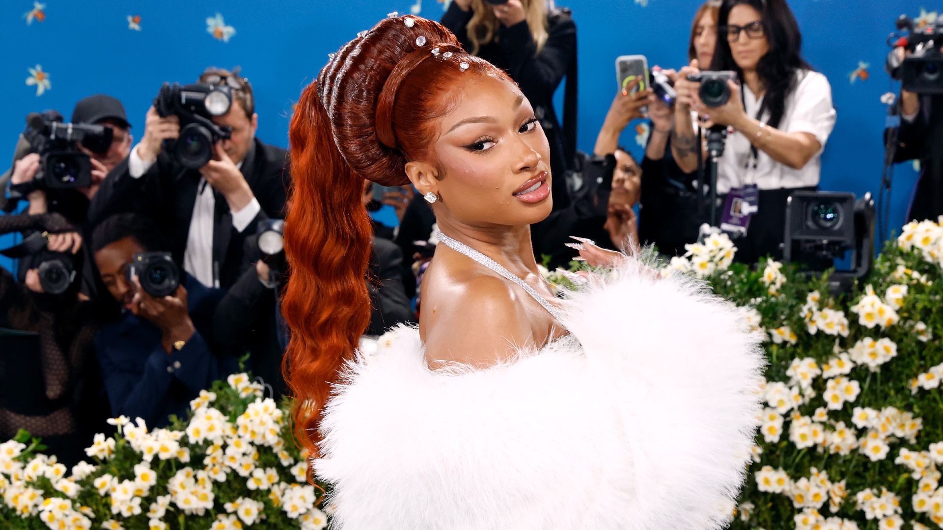 Megan Thee Stallion with red hair in an elegant white gown poses for photographers on a floral-themed carpet.