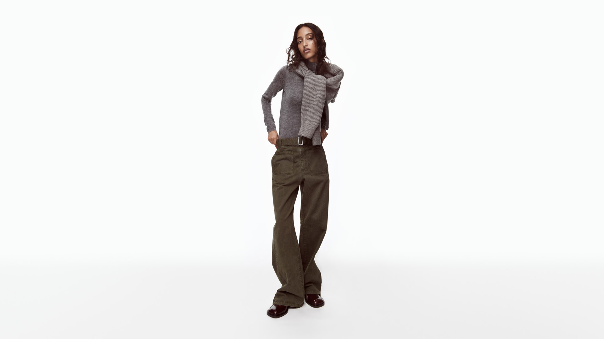 A person poses in a gray sweater, long scarf, and wide-leg pants against a plain white background.
