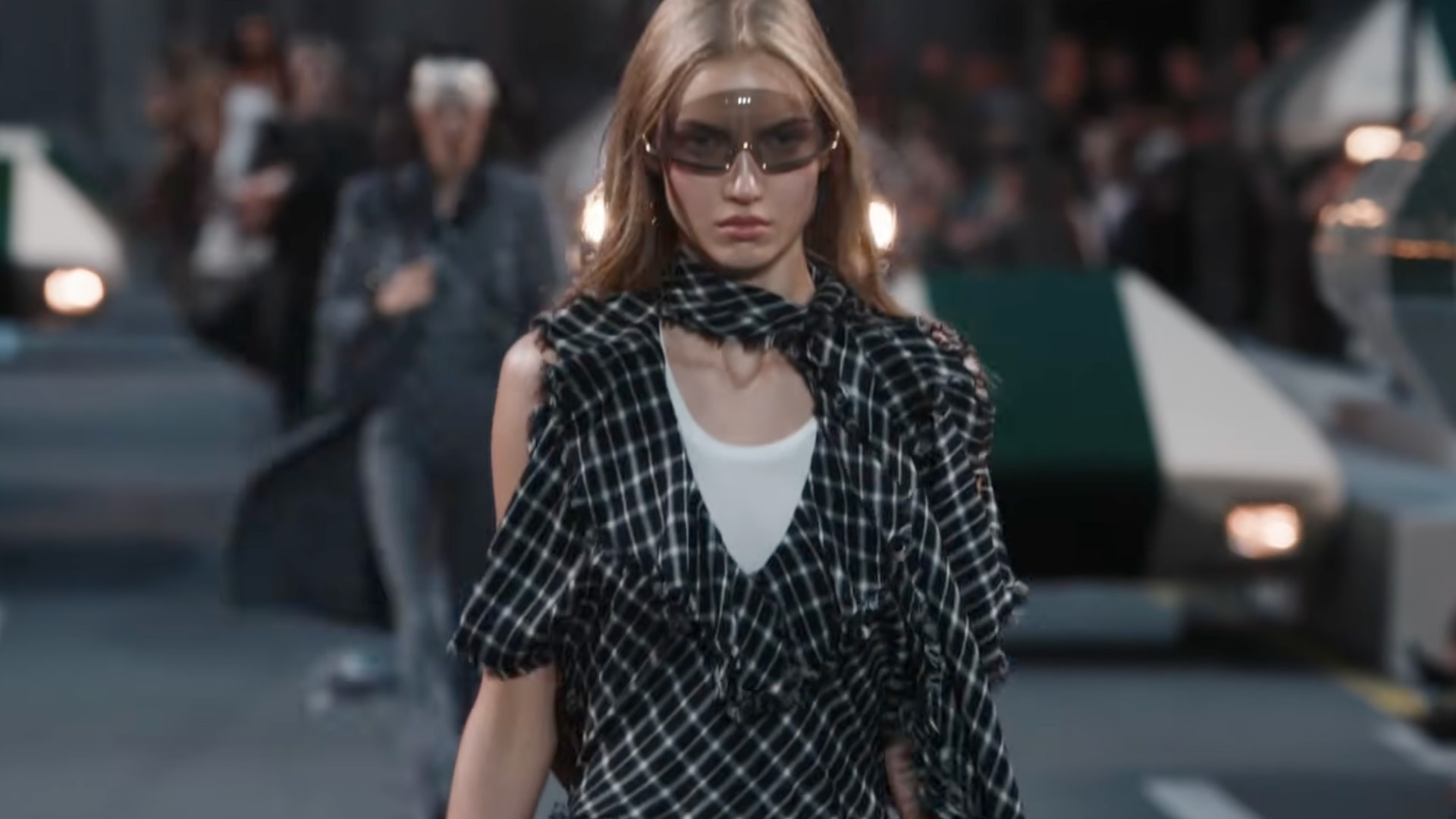 A model walks the runway wearing sunglasses and a black-and-white plaid outfit with a scarf. The background is blurred with people and lights.