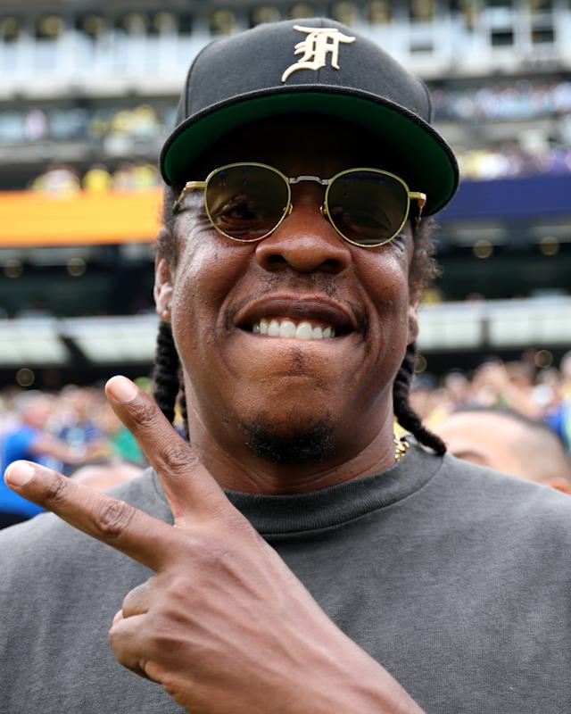 Jay-Z wearing sunglasses and a cap, making a peace sign at a sports event.