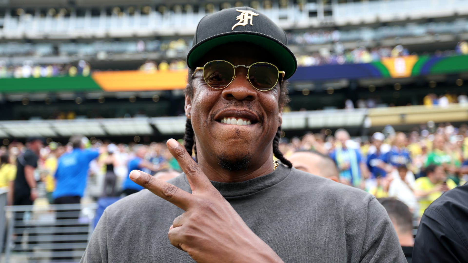 Jay-Z wearing sunglasses and a cap, making a peace sign at a sports event.