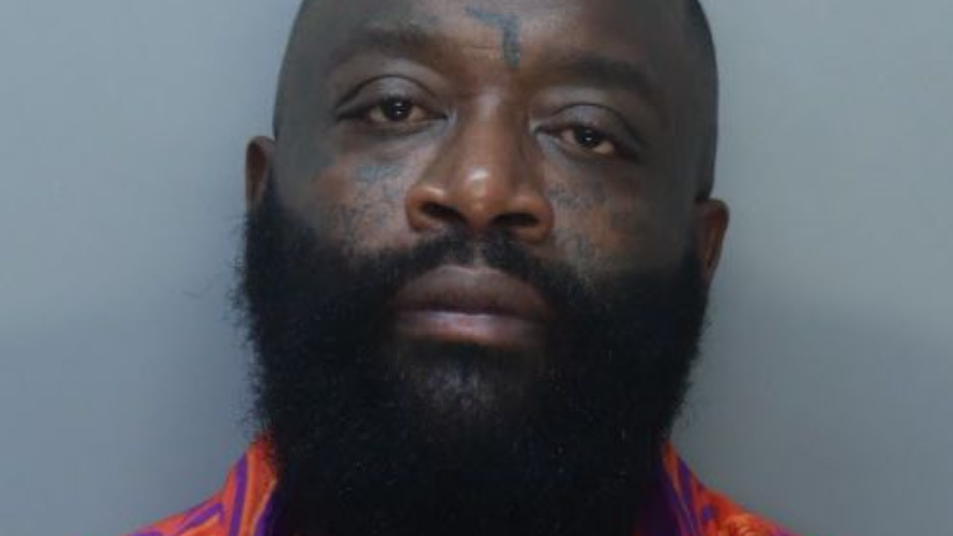 A man with a beard and a facial tattoo is wearing a colorful shirt, looking directly at the camera with a neutral expression.