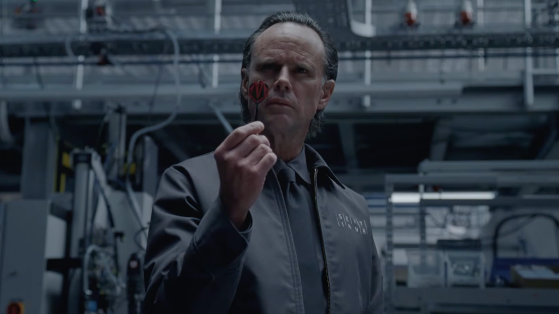 Walton Goggins in a gray jacket holds a small object in a futuristic industrial setting.