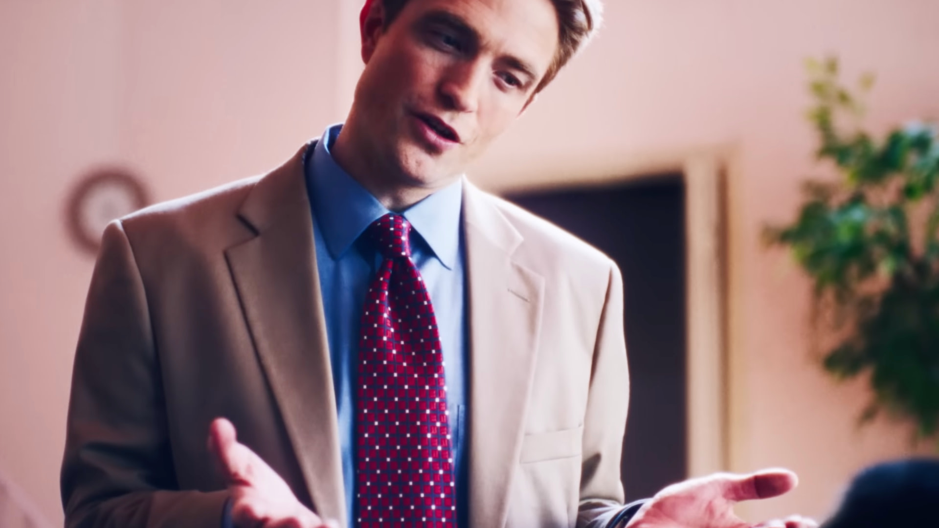 Robert Pattinson in a beige suit and red polka dot tie gestures while speaking, with a blurred background and a plant visible.