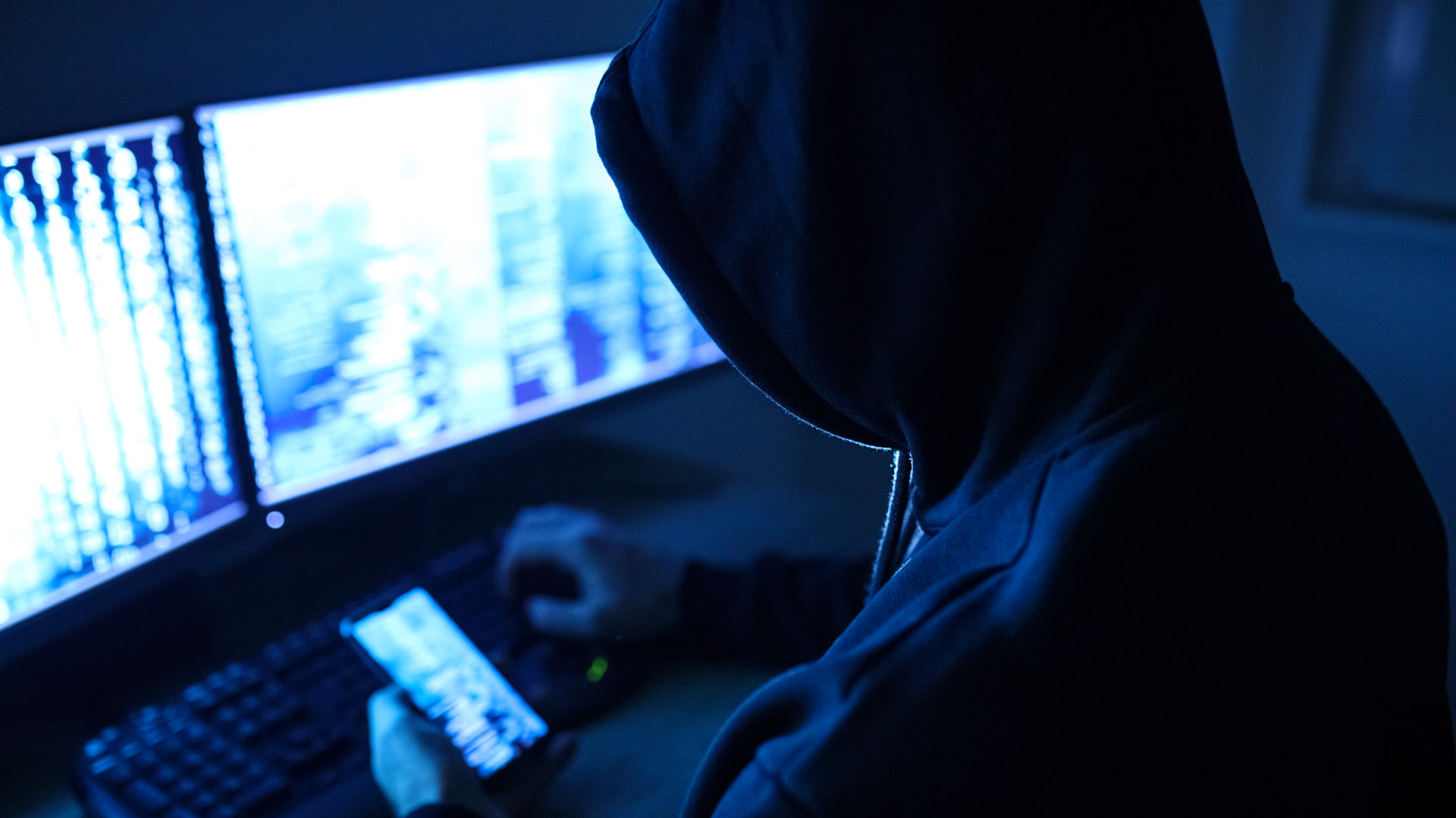 A person in a dark hoodie sits at a computer with multiple monitors, holding a smartphone, in a dimly lit room.