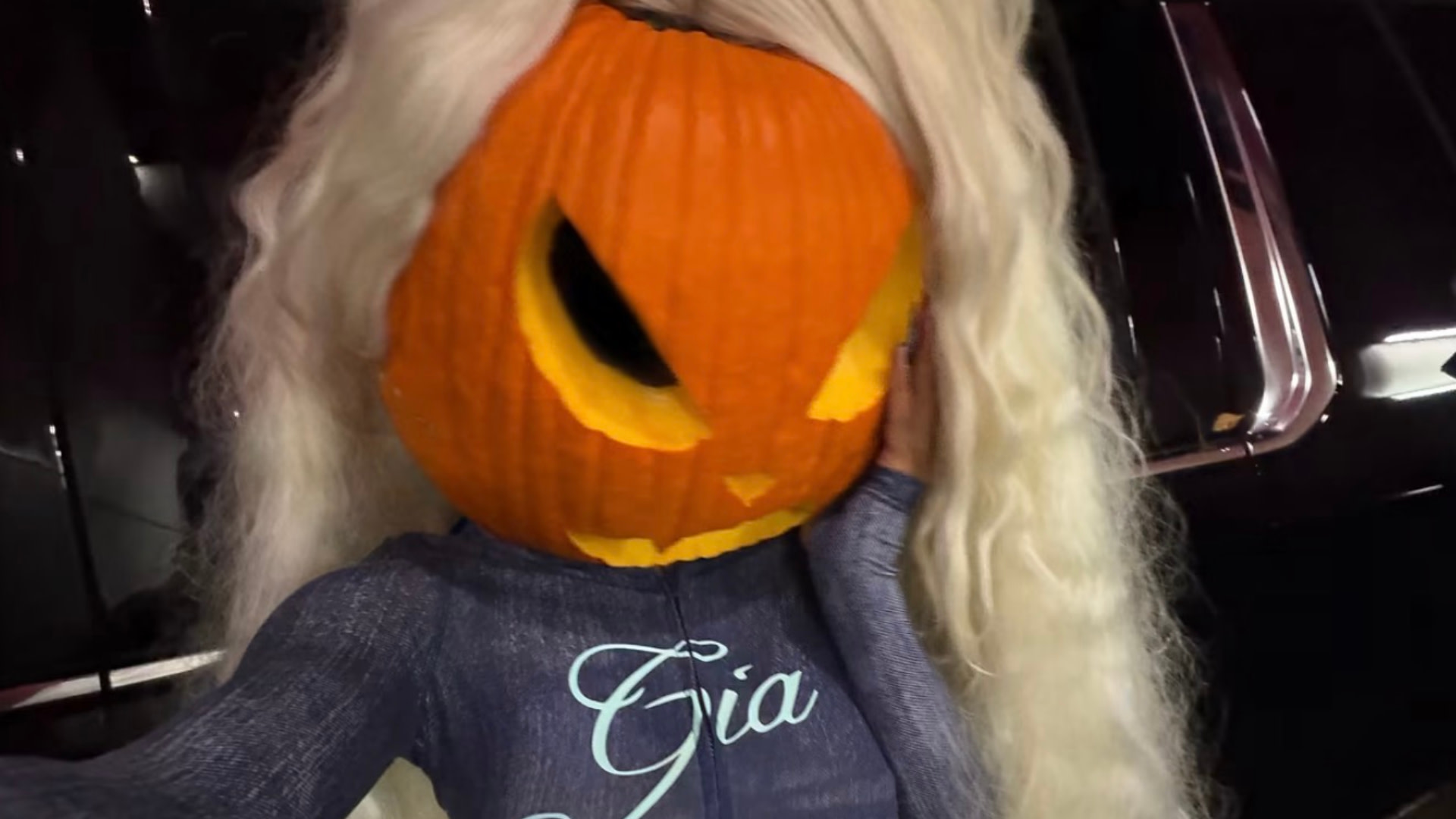 Megan Thee Stallion with a pumpkin carved as a jack-o'-lantern on their head, wearing a blue outfit with "Gia" on it, and long blonde hair.