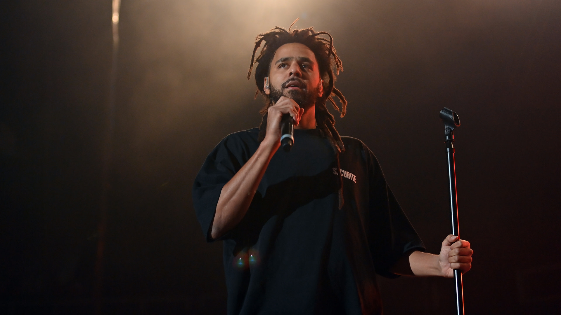 J. Cole performing on stage, holding a microphone, with dramatic lighting.