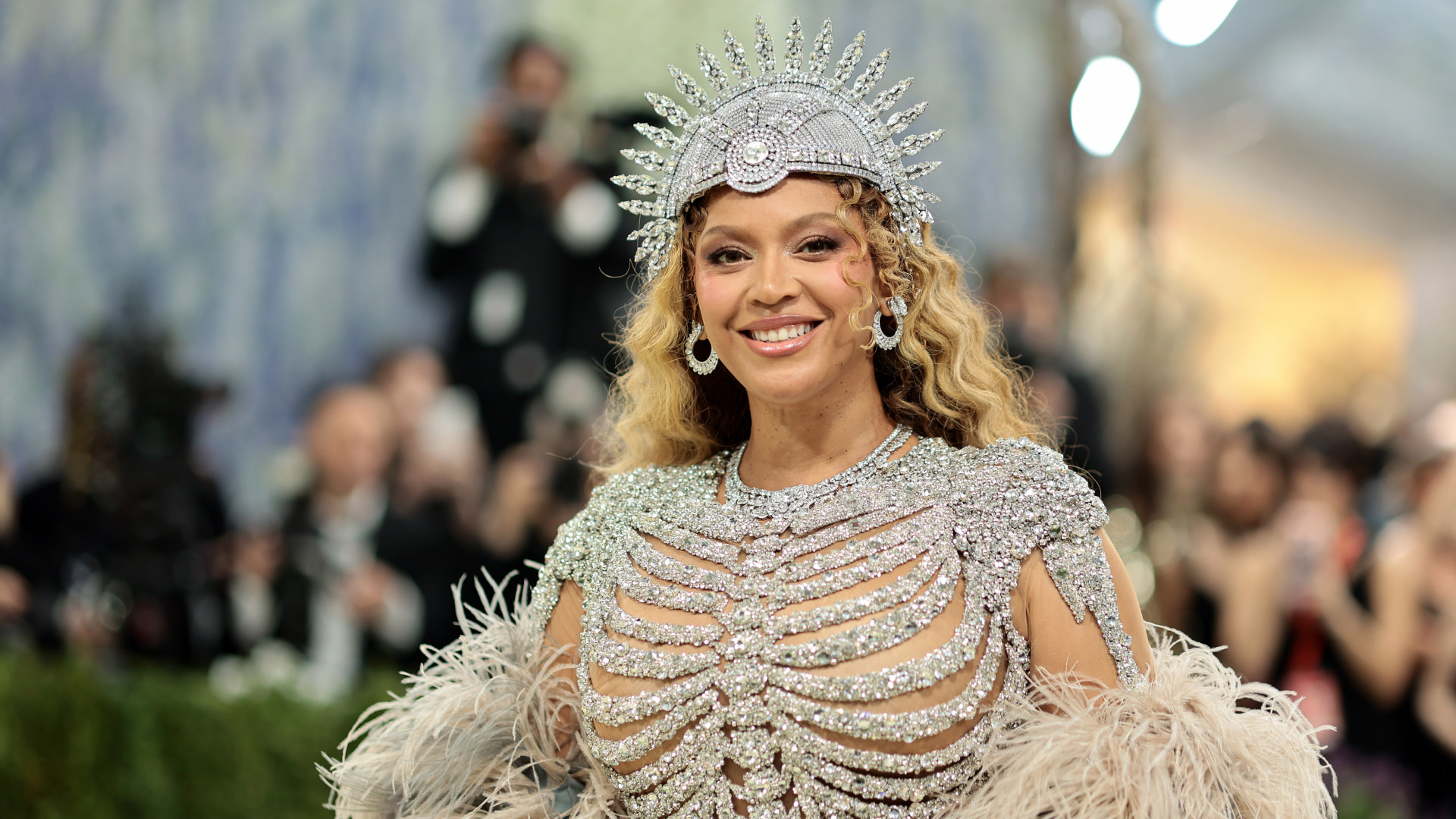 Beyoncé in a sparkling, ornate outfit with a feathered design and a jeweled headdress, smiling at an event.