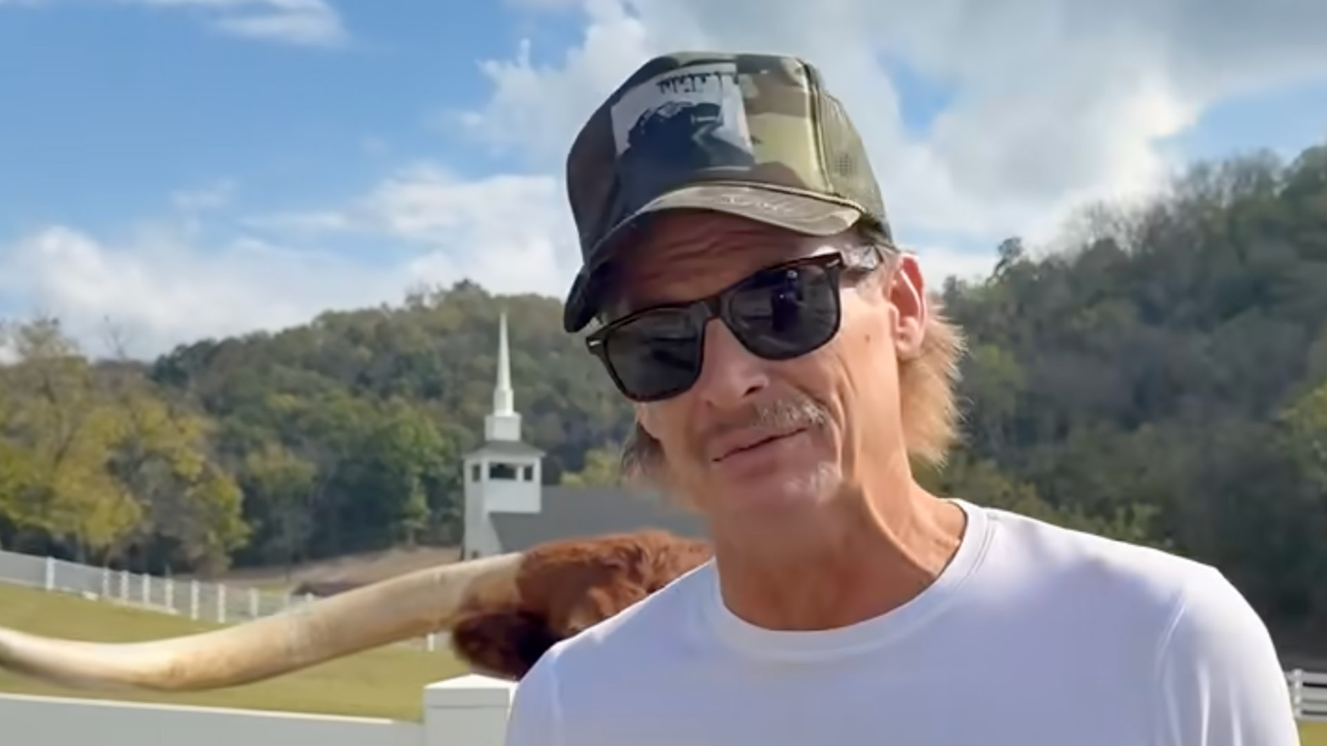 Kid Rock wearing sunglasses and a camo cap stands outdoors, with a longhorn in the background and a small church visible behind.
